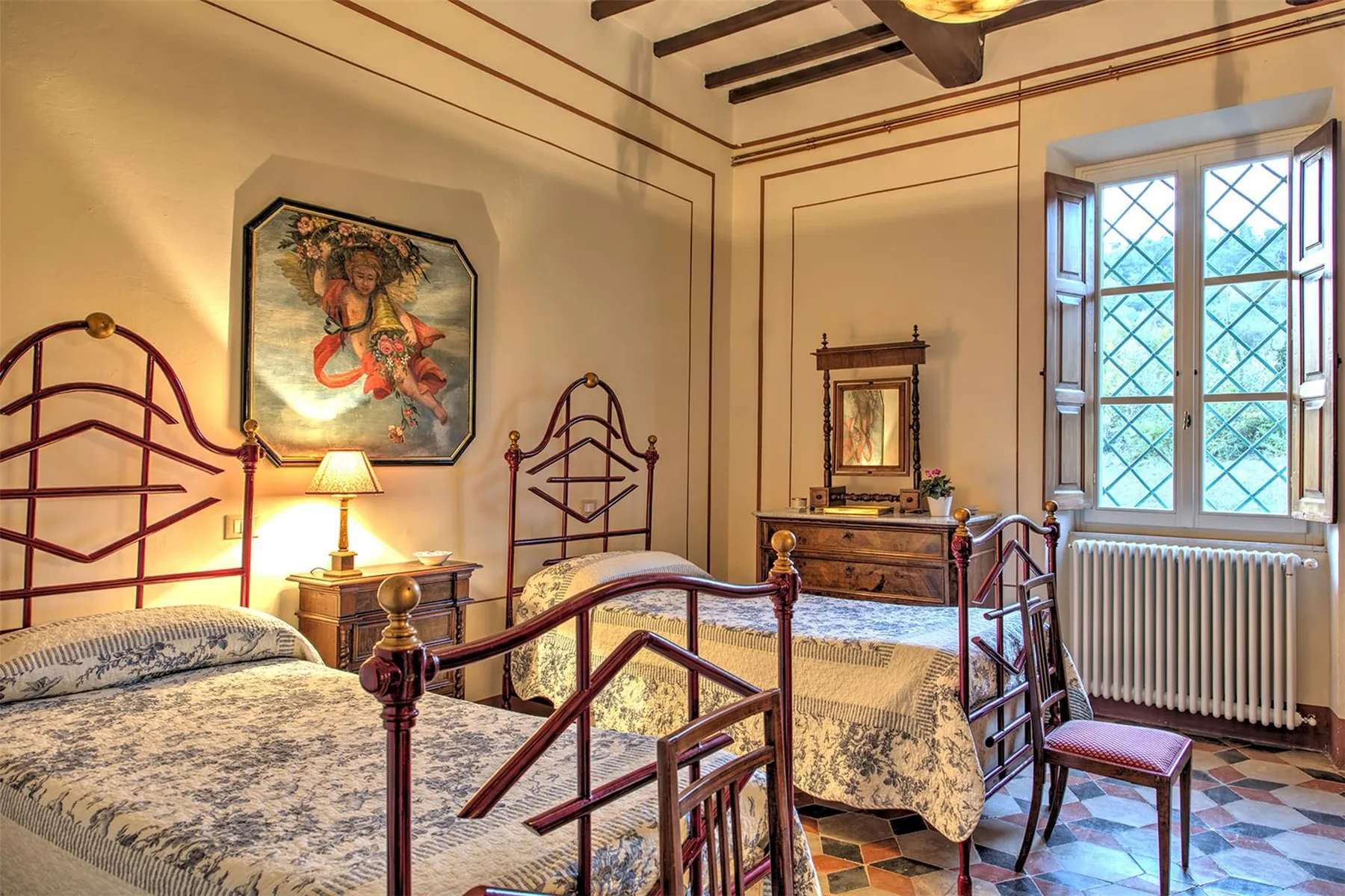 Un dormitorio tradicional con dos camas de armazón metálico, ropa de cama estampada, una cómoda de madera y una ventana grande.