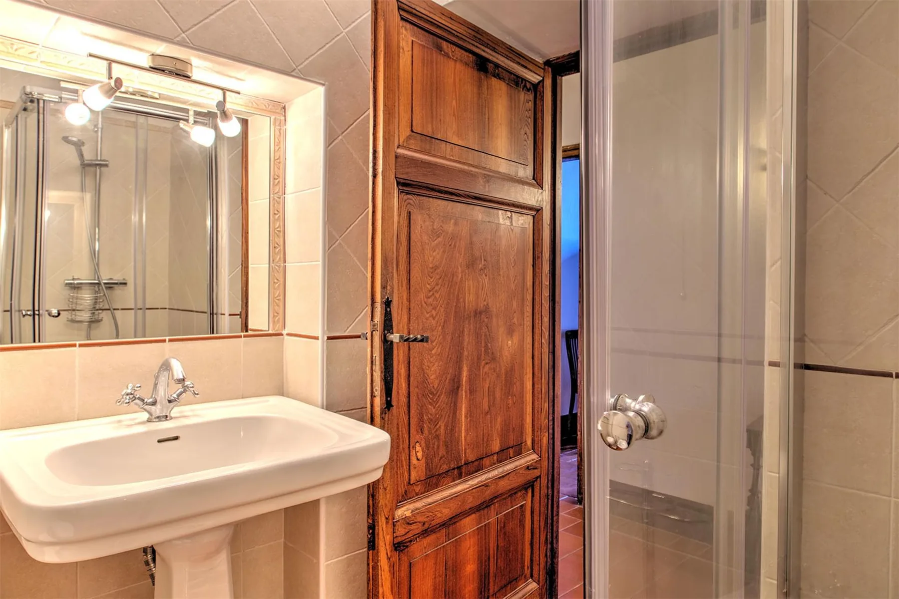 Baño alicatado con lavabo blanco de pedestal, espejo de pared, ducha con mampara de vidrio y puerta de madera.
