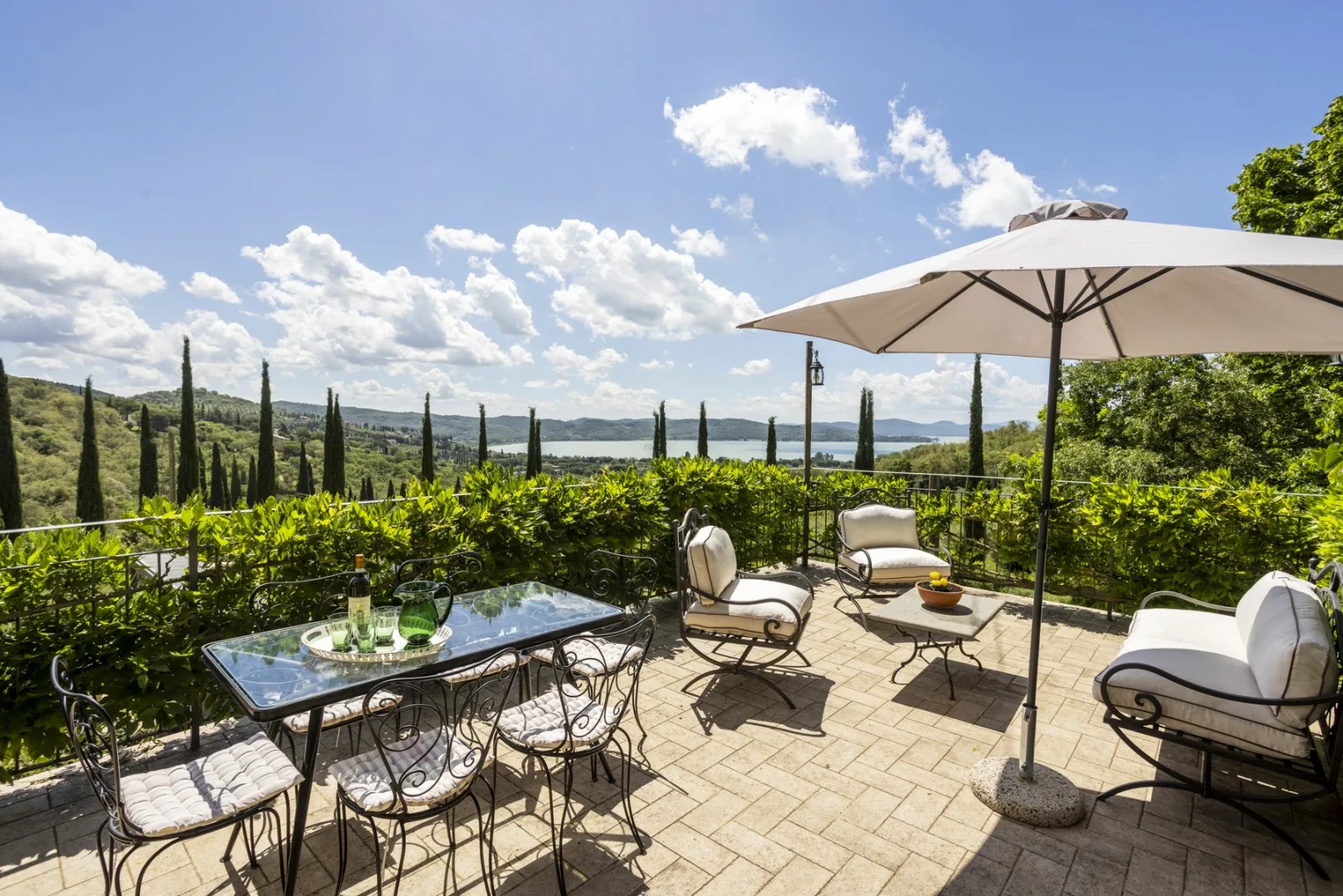 Una terraza soleada con mesa de comedor, sillas con cojines y una sombrilla ofrece vistas a un viñedo y colinas lejanas.