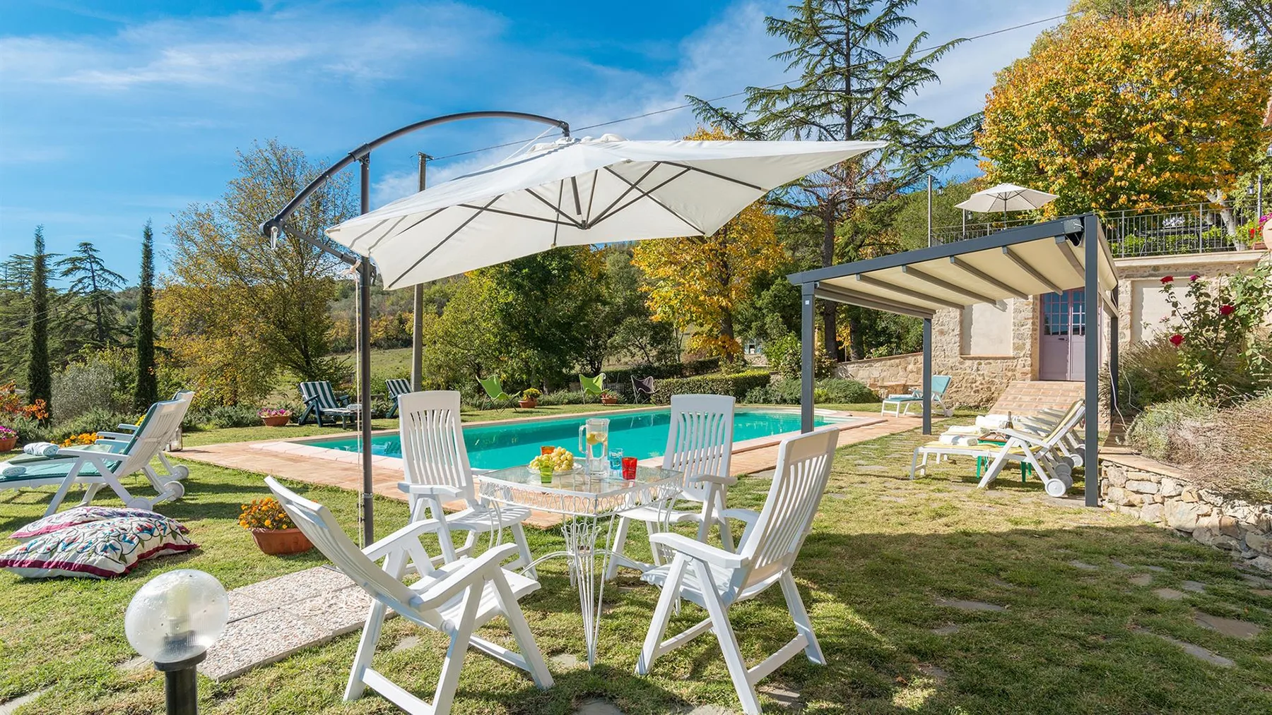 Un patio ajardinado con sillas blancas y una mesa está junto a una piscina turquesa.