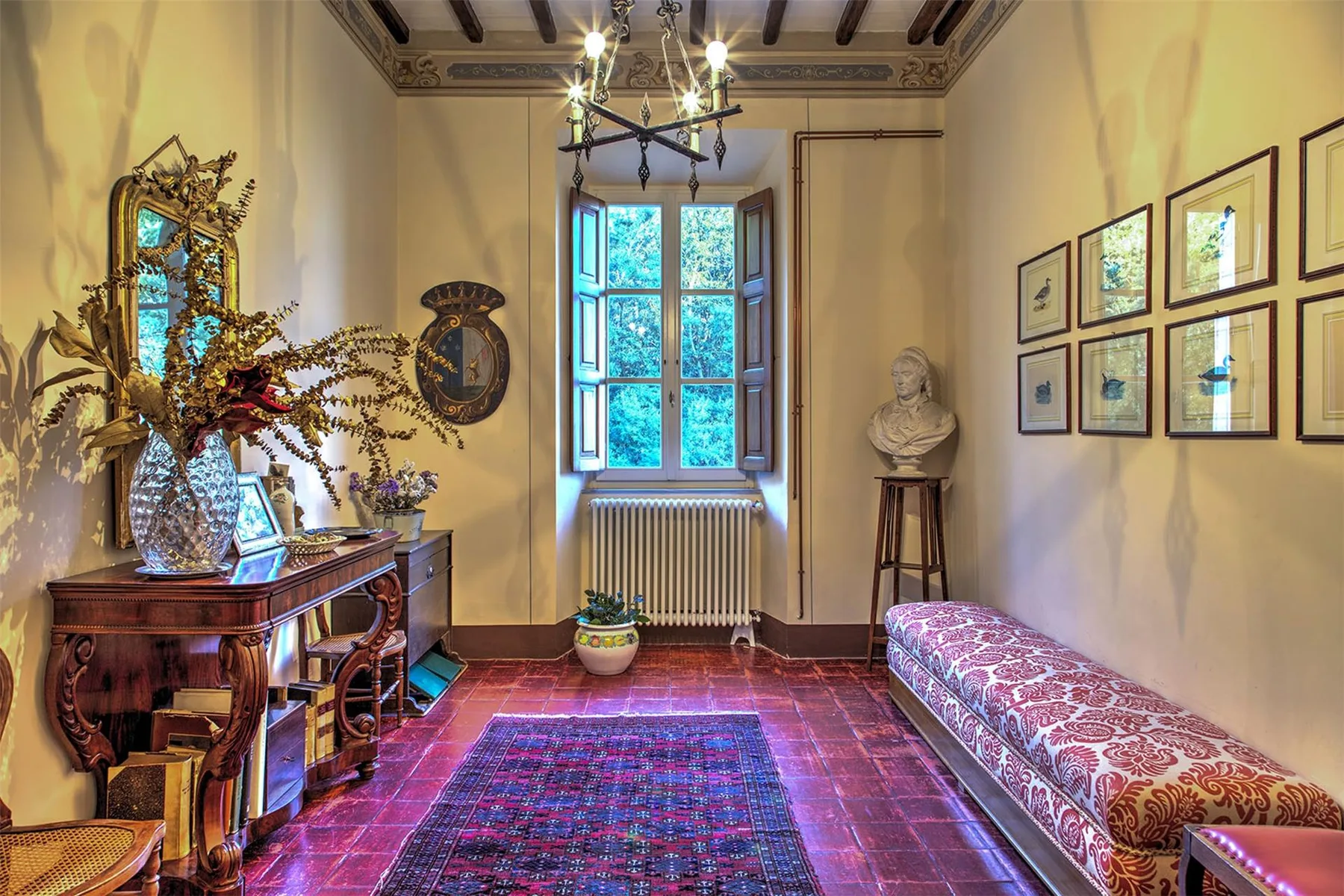 Una sala amueblada tiene una alfombra roja estampada, una ventana con radiador y un largo banco tapizado.