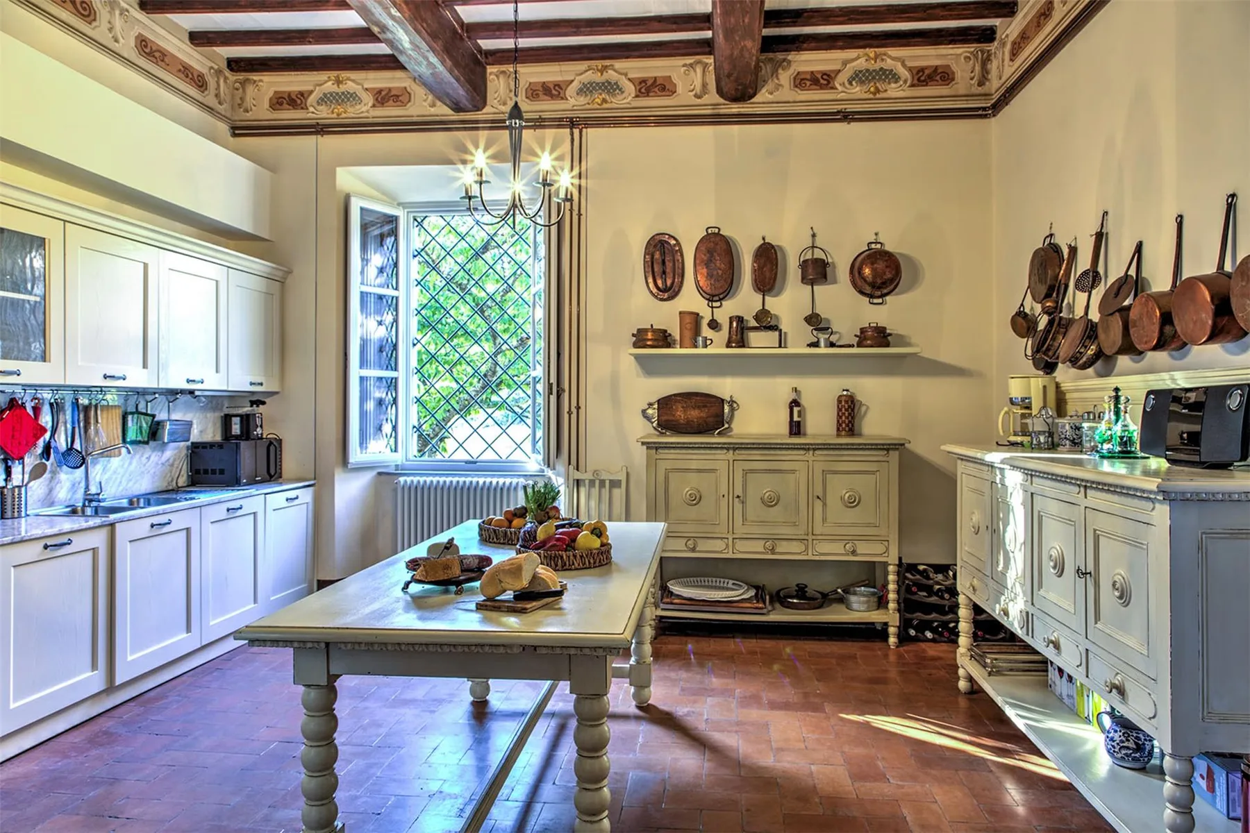 Una cocina amplia cuenta con una mesa central de madera, armarios blancos, utensilios colgados y un techo decorado con vigas.