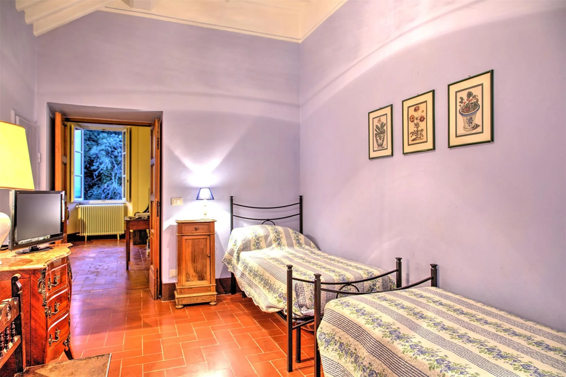 Un dormitorio con dos camas de hierro, una cómoda de madera y láminas botánicas en paredes moradas claras.