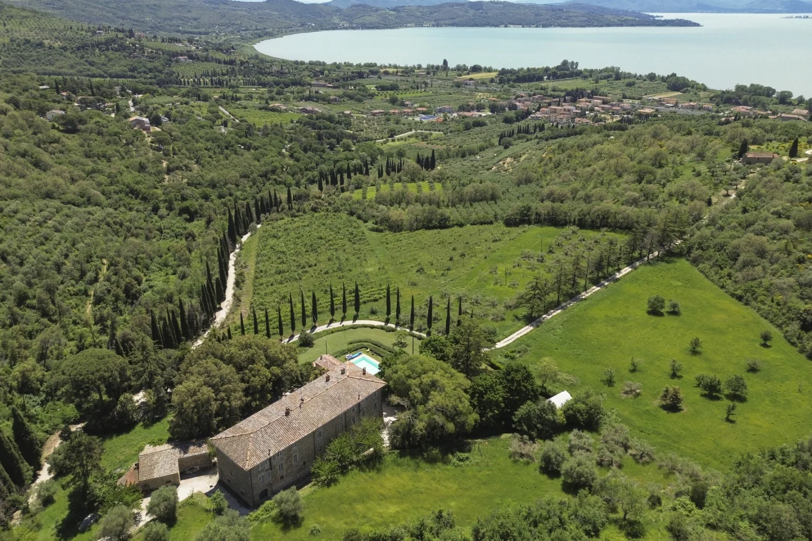 Una vista aérea muestra un edificio de piedra entre campos verdes, caminos arbolados y un lago a lo lejos.