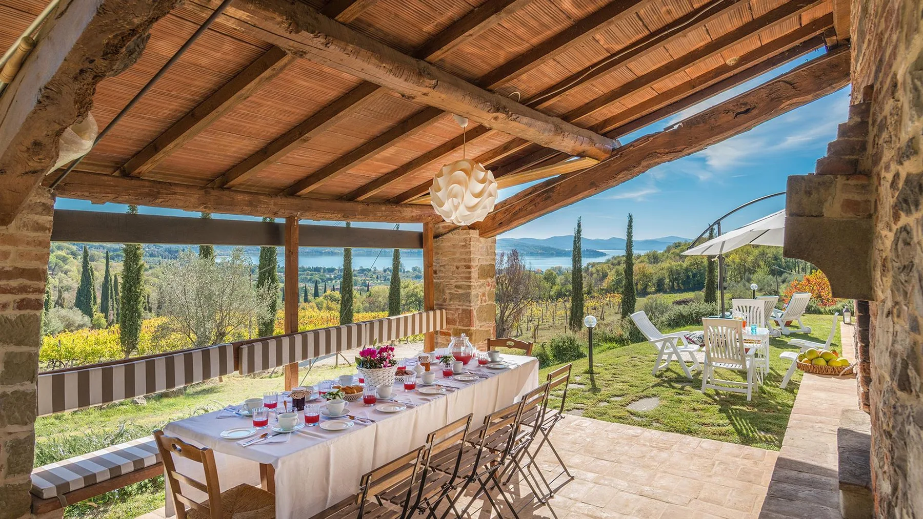 Una zona cubierta para comer al aire libre tiene una mesa larga preparada, sillas de madera y vistas al jardín y las colinas.