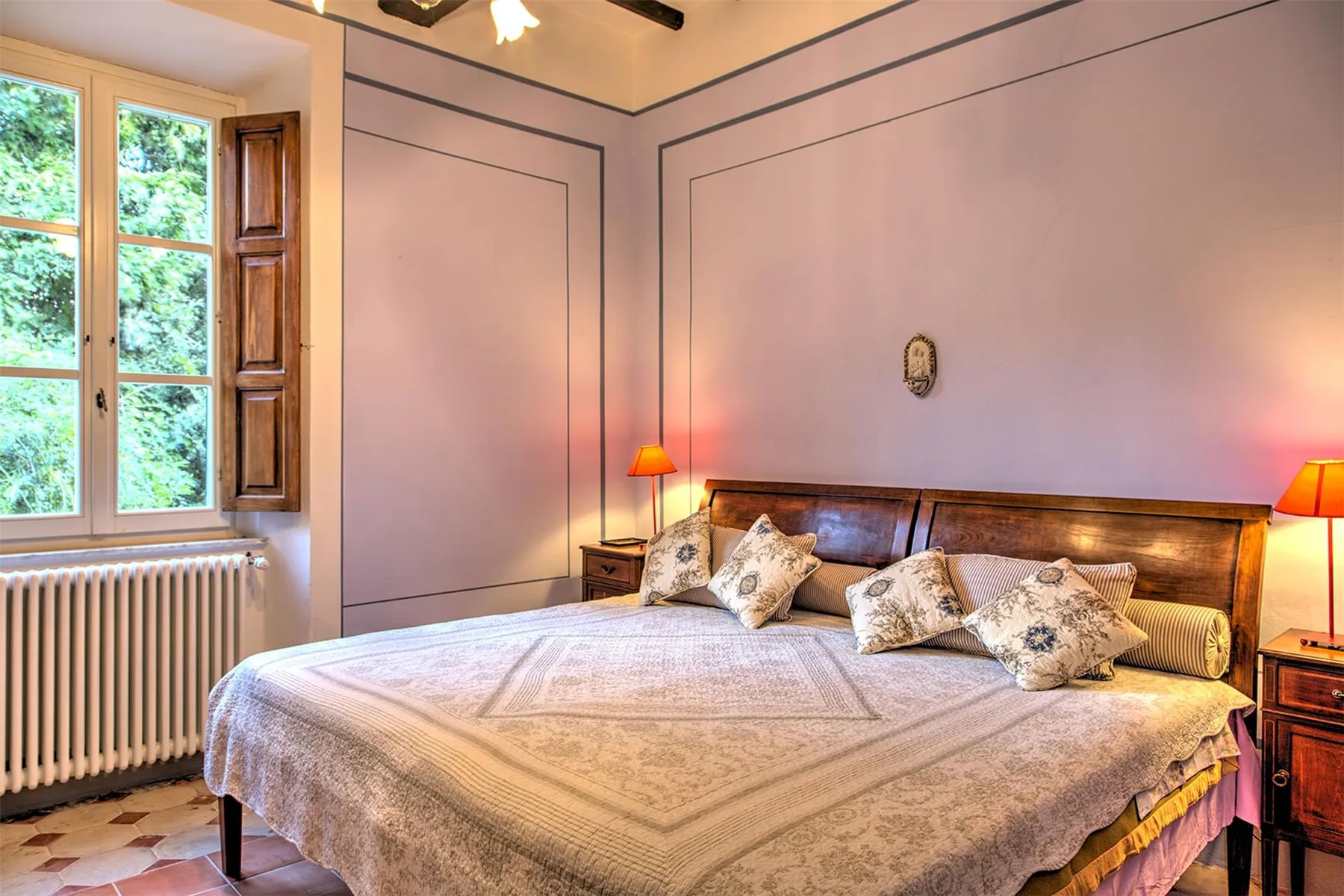 Un dormitorio con una cama grande, ropa de cama estampada, lámparas de noche y una ventana alta con vistas a la vegetación.