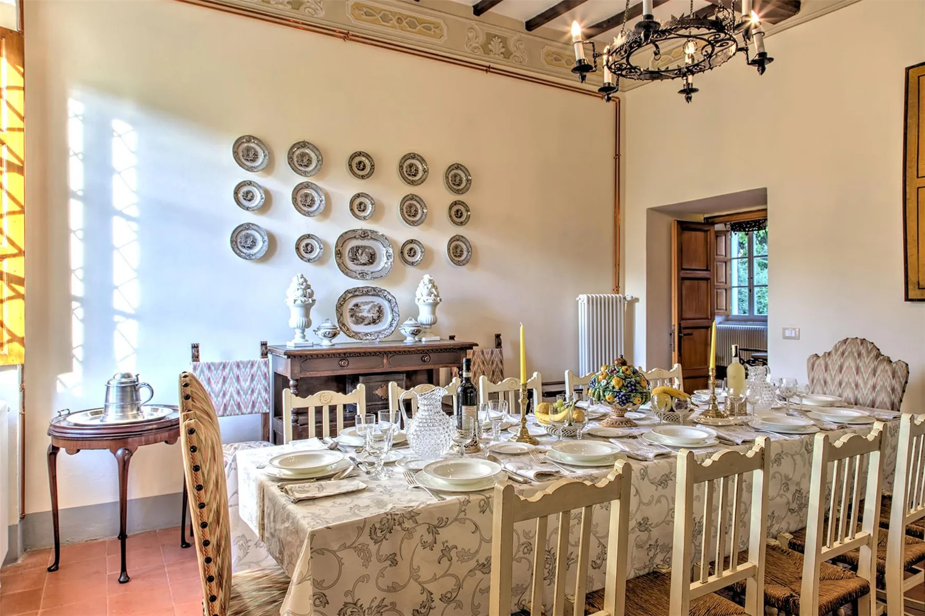 Un comedor formal tiene una mesa larga puesta, sillas talladas, platos decorativos y una gran lámpara de araña.