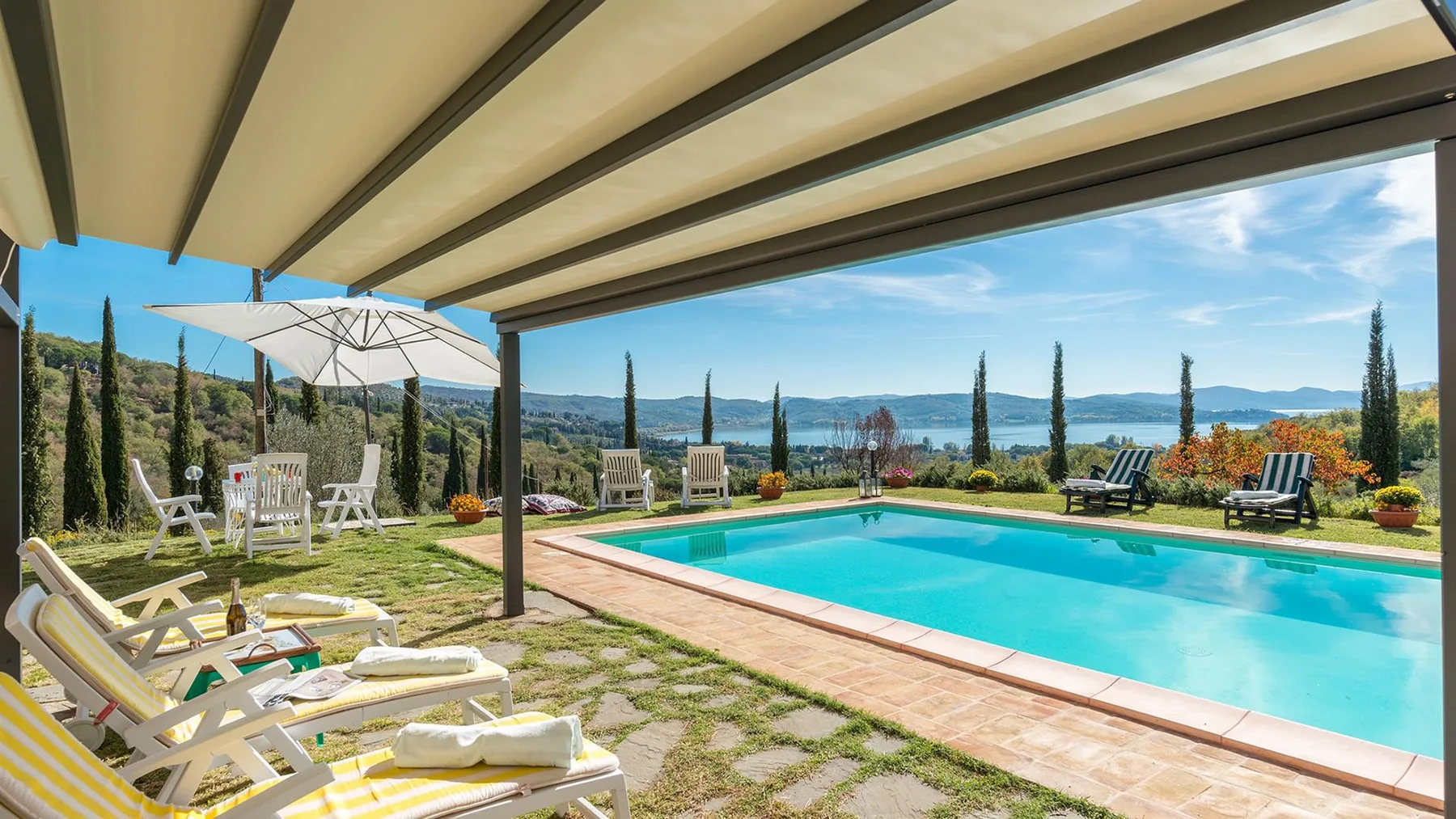 Una piscina turquesa está junto a un patio de piedra con tumbonas, bajo una pérgola sombreada con vistas a las colinas.