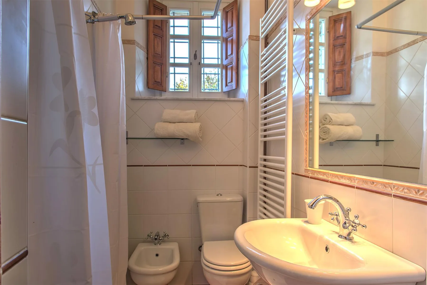 Un baño alicatado tiene bañera, inodoro, bidé, toallero y un lavabo bajo un espejo grande.