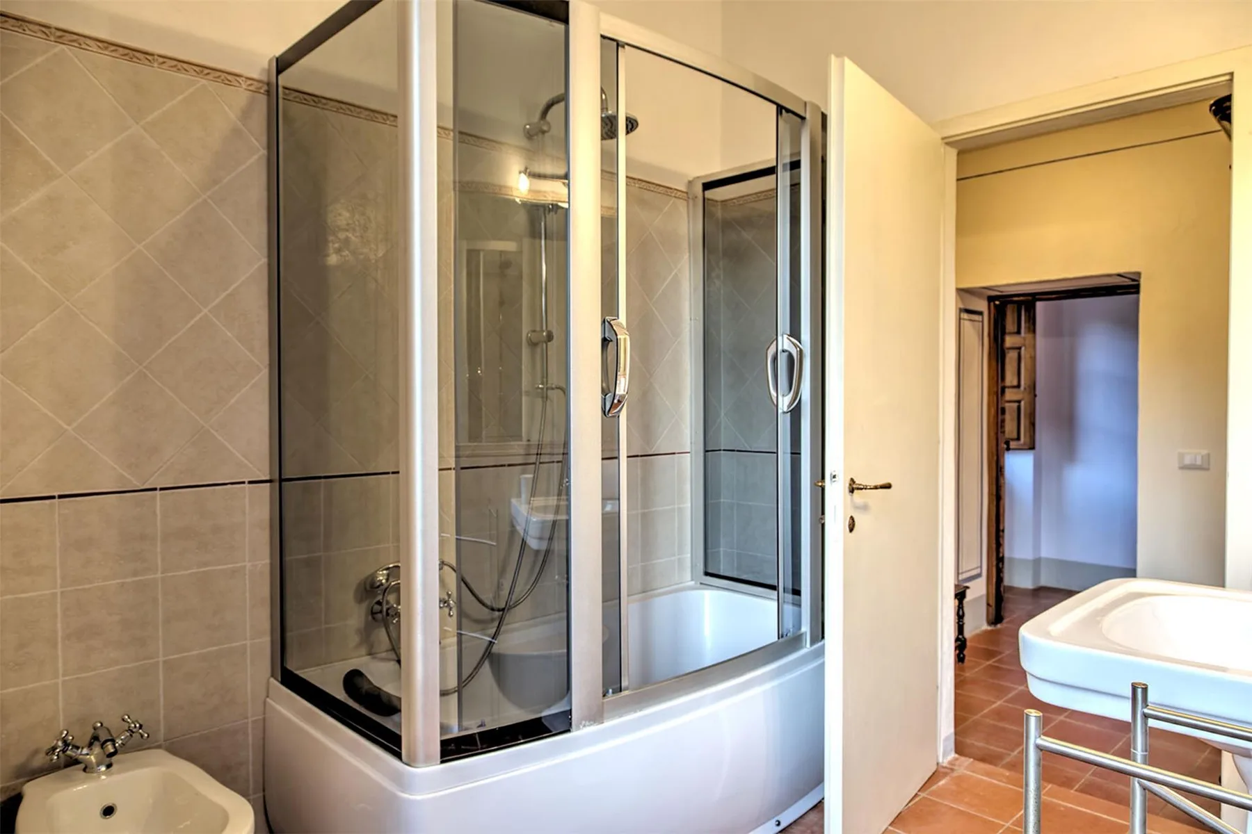 Un baño tiene una bañera con ducha y mampara de vidrio, un inodoro y un lavabo blanco junto a una puerta abierta.