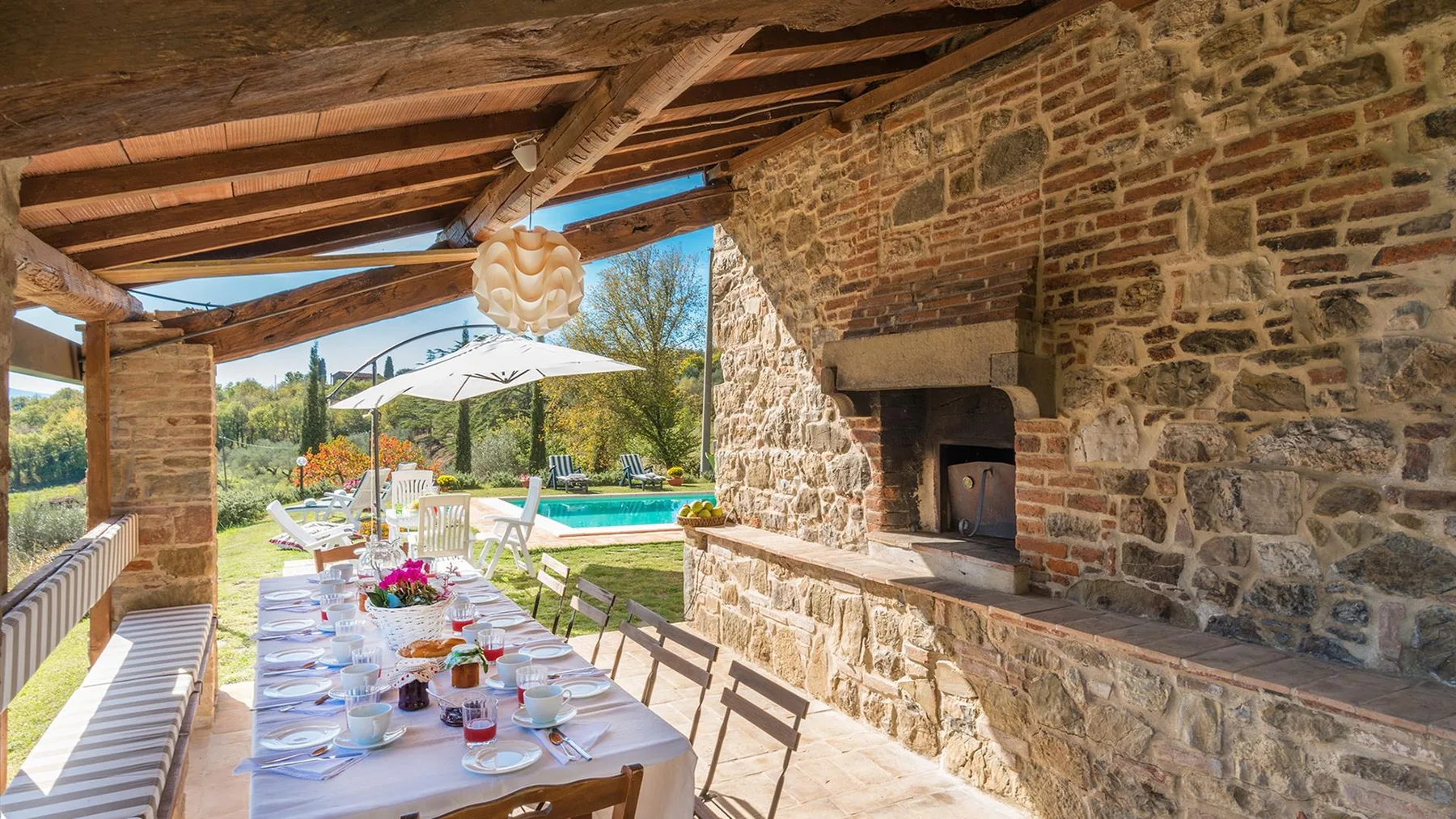 Un patio cubierto de piedra tiene una larga mesa puesta junto a una chimenea empotrada, con vistas a un jardín y una piscina.