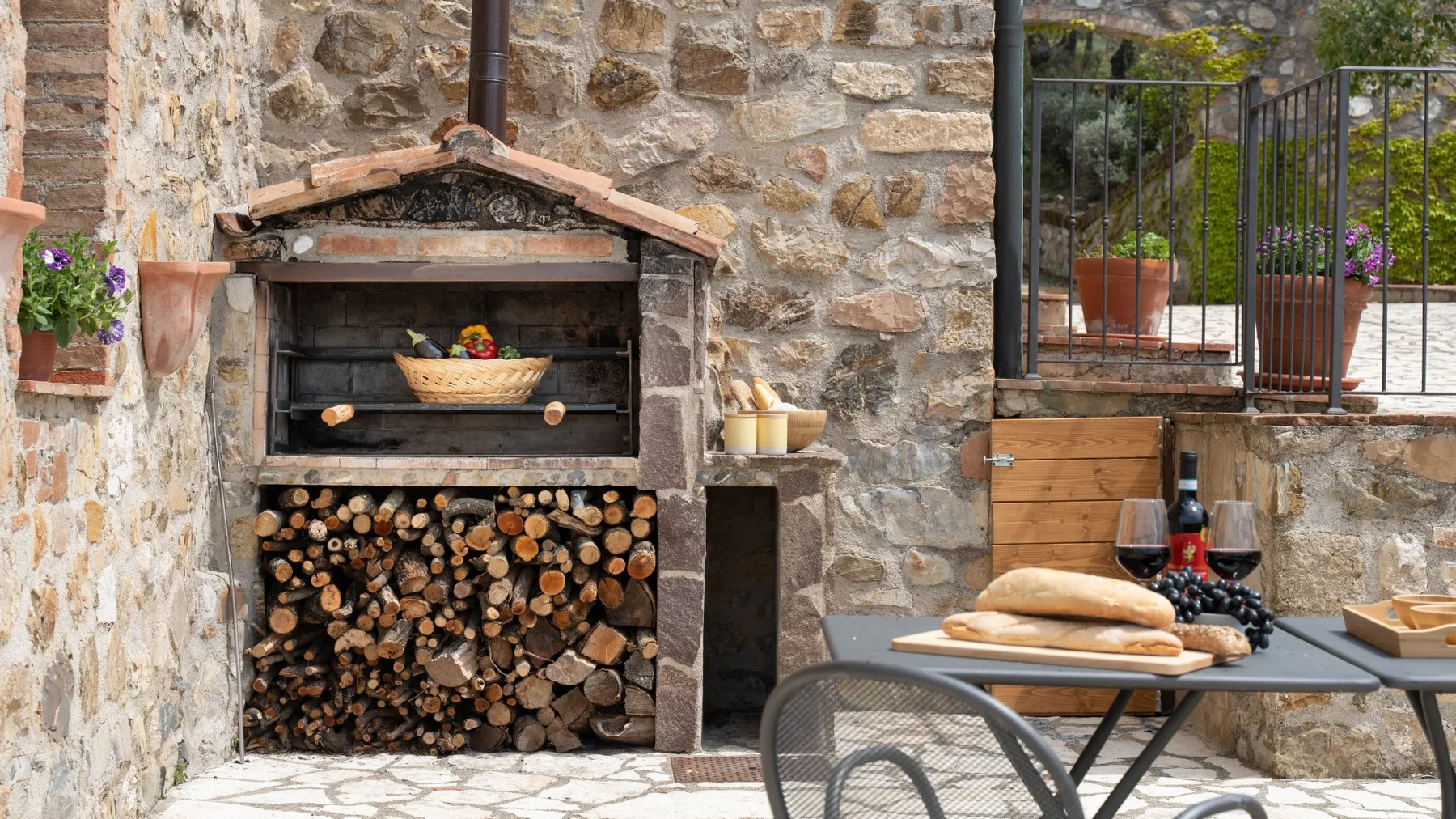 Un comedor rústico al aire libre cuenta con una chimenea de piedra y una mesa con pan y vino.