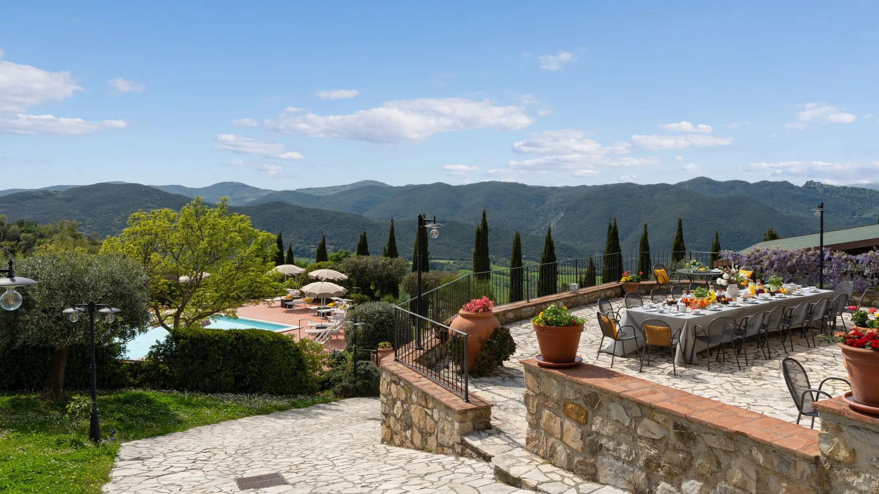 Una terraza de piedra mira hacia una piscina, un comedor al aire libre y colinas lejanas.