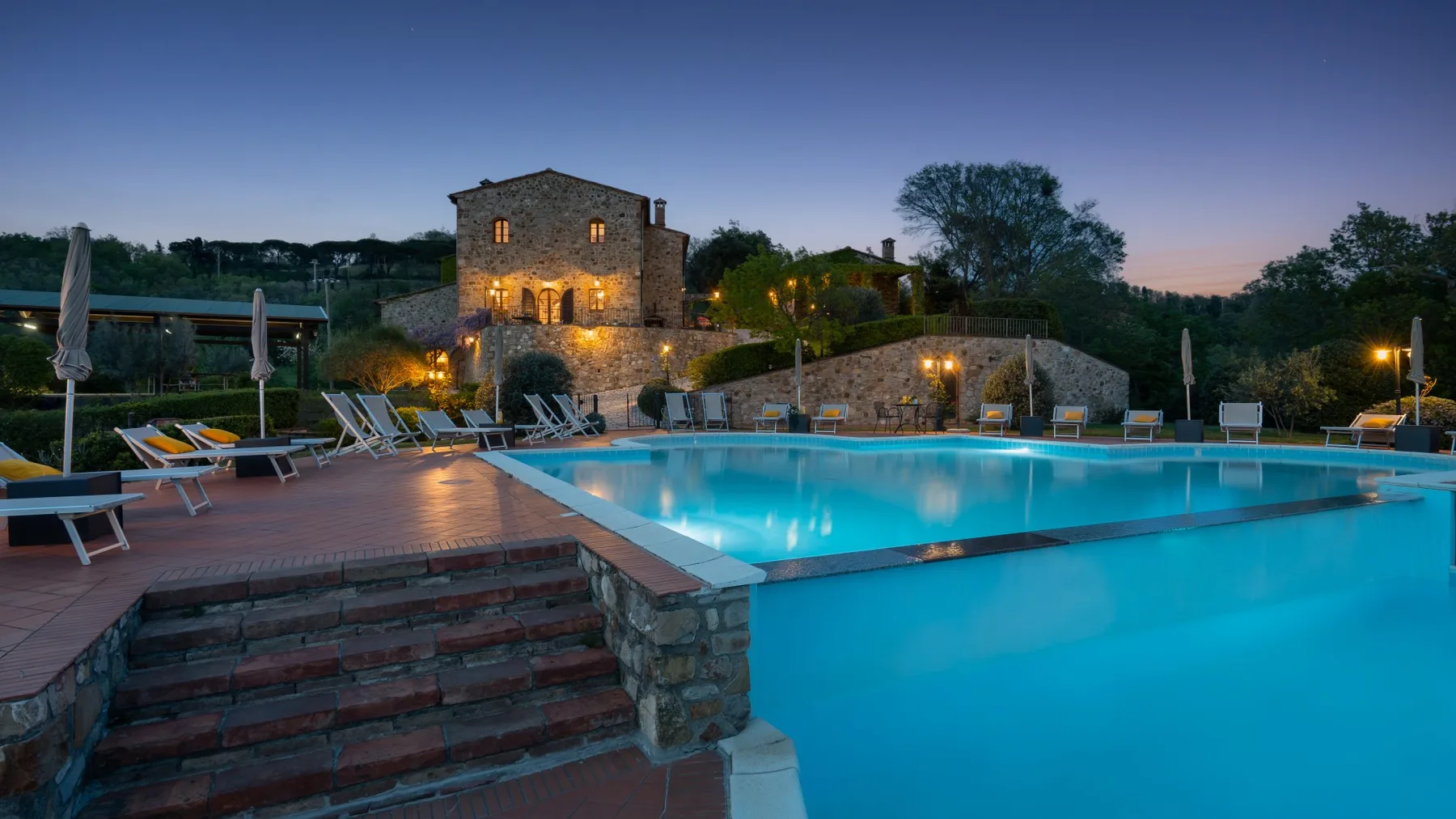 Una piscina exterior iluminada junto a un edificio de piedra al anochecer.