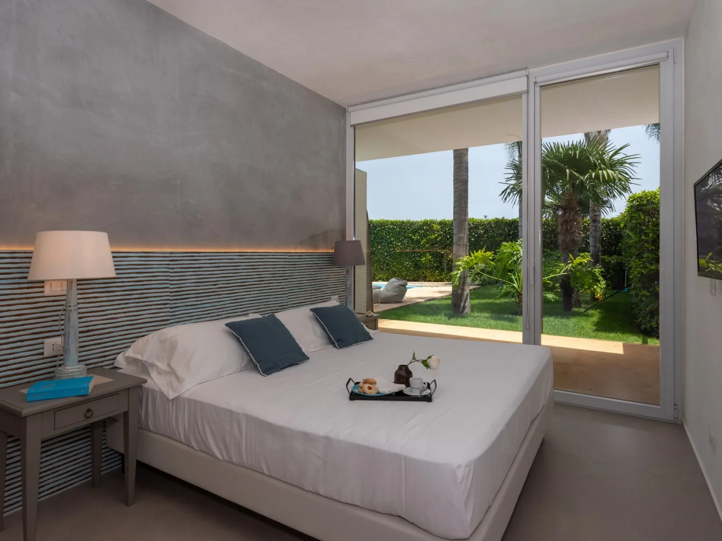 A bedroom with a neatly made bed beside large windows overlooking a garden.