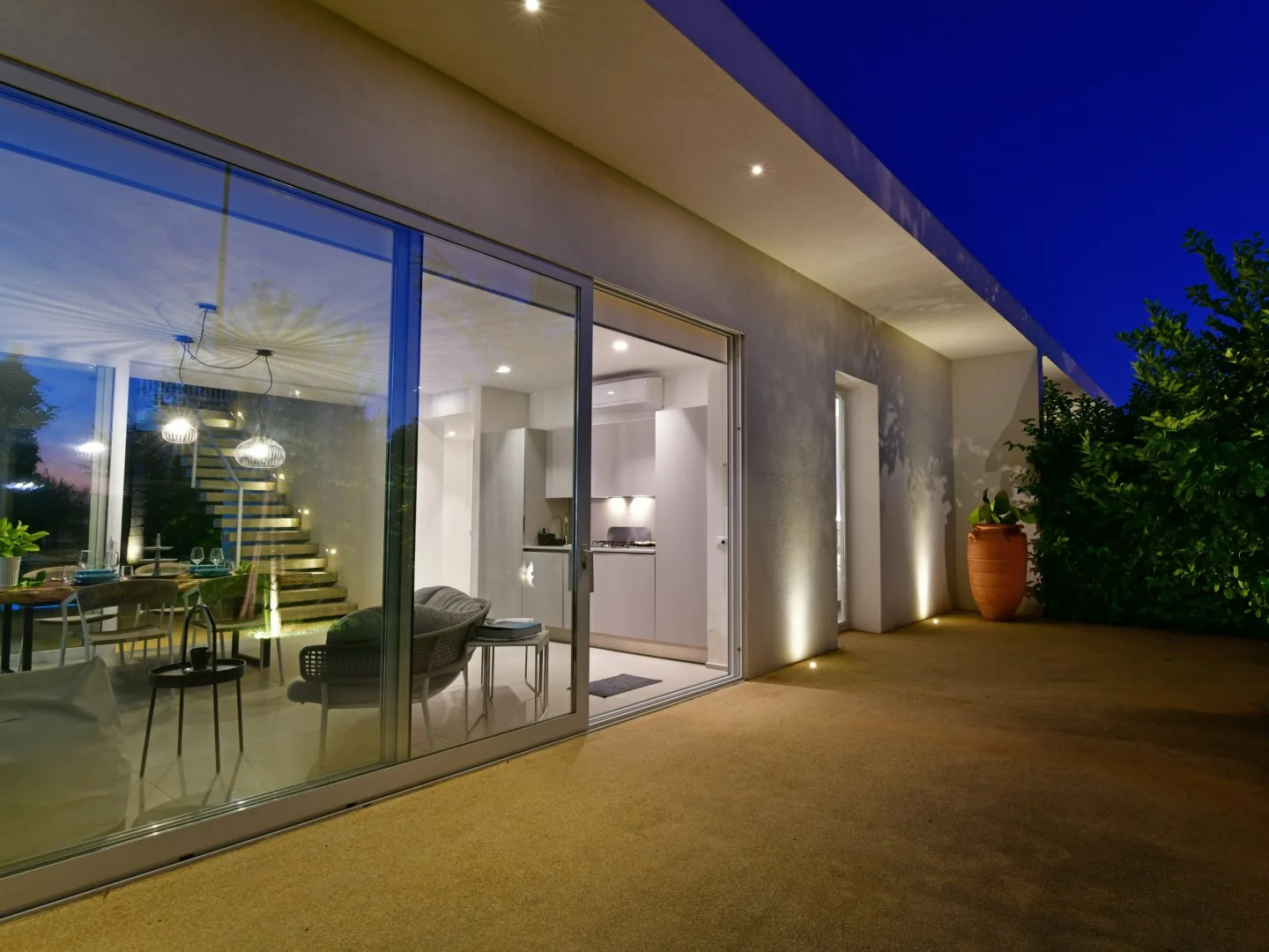 A modern house exterior at dusk, with large glass doors opening onto a lit interior.