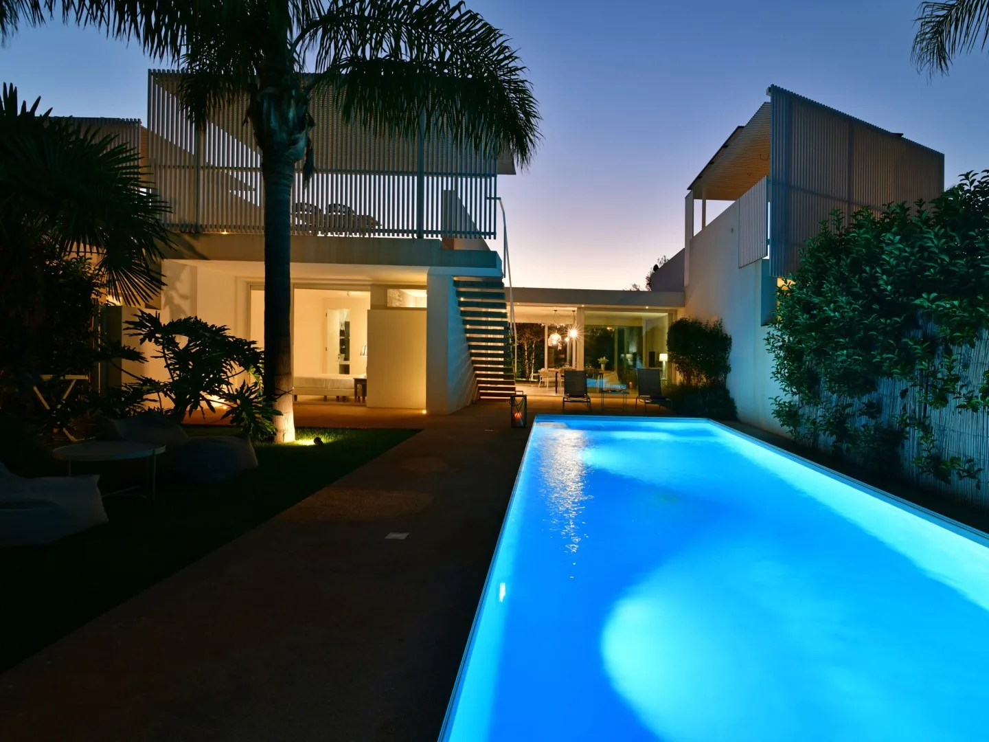 A modern house and illuminated swimming pool at dusk, surrounded by palm trees.