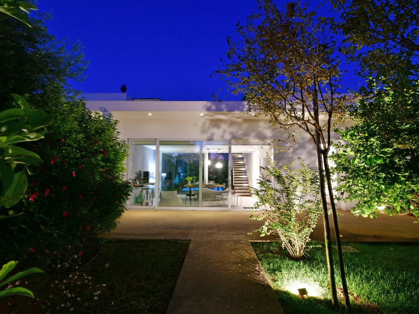 A modern house glows at dusk behind a garden path and leafy landscaping.