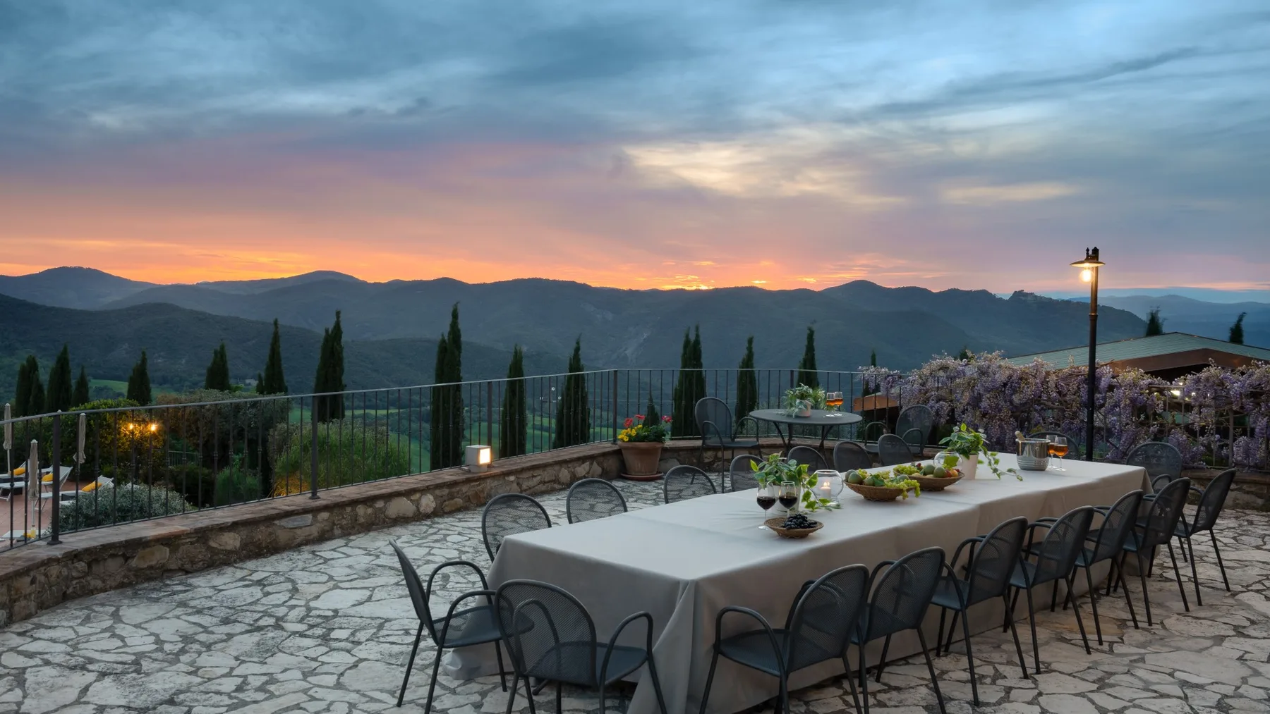 Una terraza de comedor al aire libre mira hacia montañas bajo un cielo de atardecer colorido.