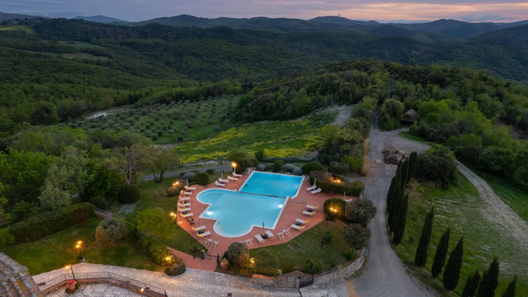 Una piscina al aire libre y asientos alrededor se encuentran entre colinas boscosas al atardecer.