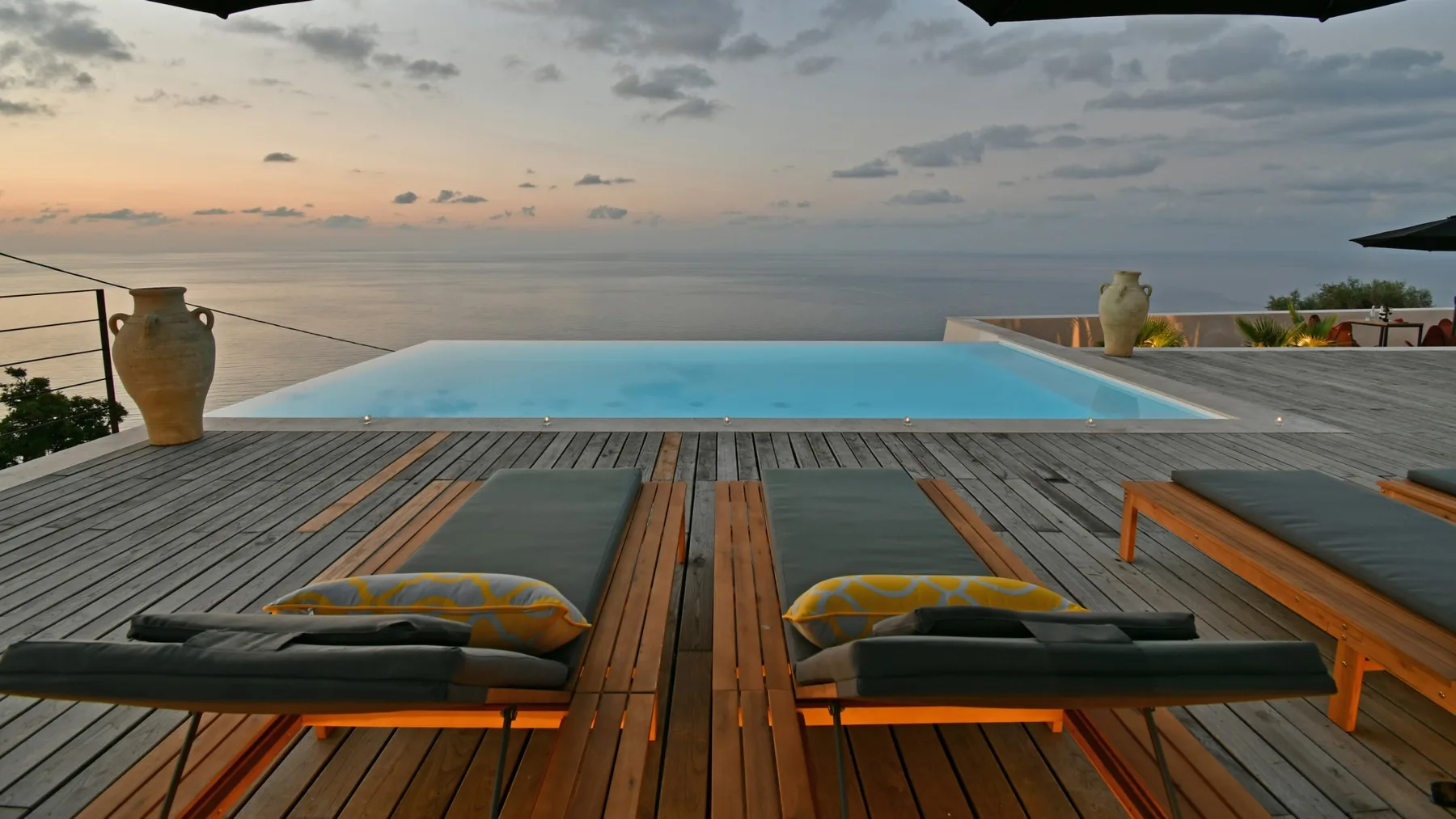 Una piscina infinita da al océano al atardecer, con una terraza de madera y tumbonas acolchadas en primer plano.