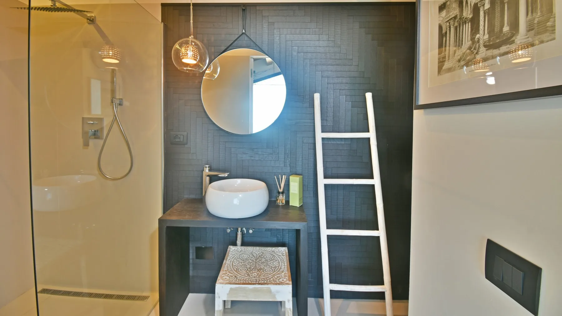 Un baño moderno cuenta con ducha de vidrio, lavabo sobre encimera, espejo redondo y una escalera blanca sobre azulejos oscuros.
