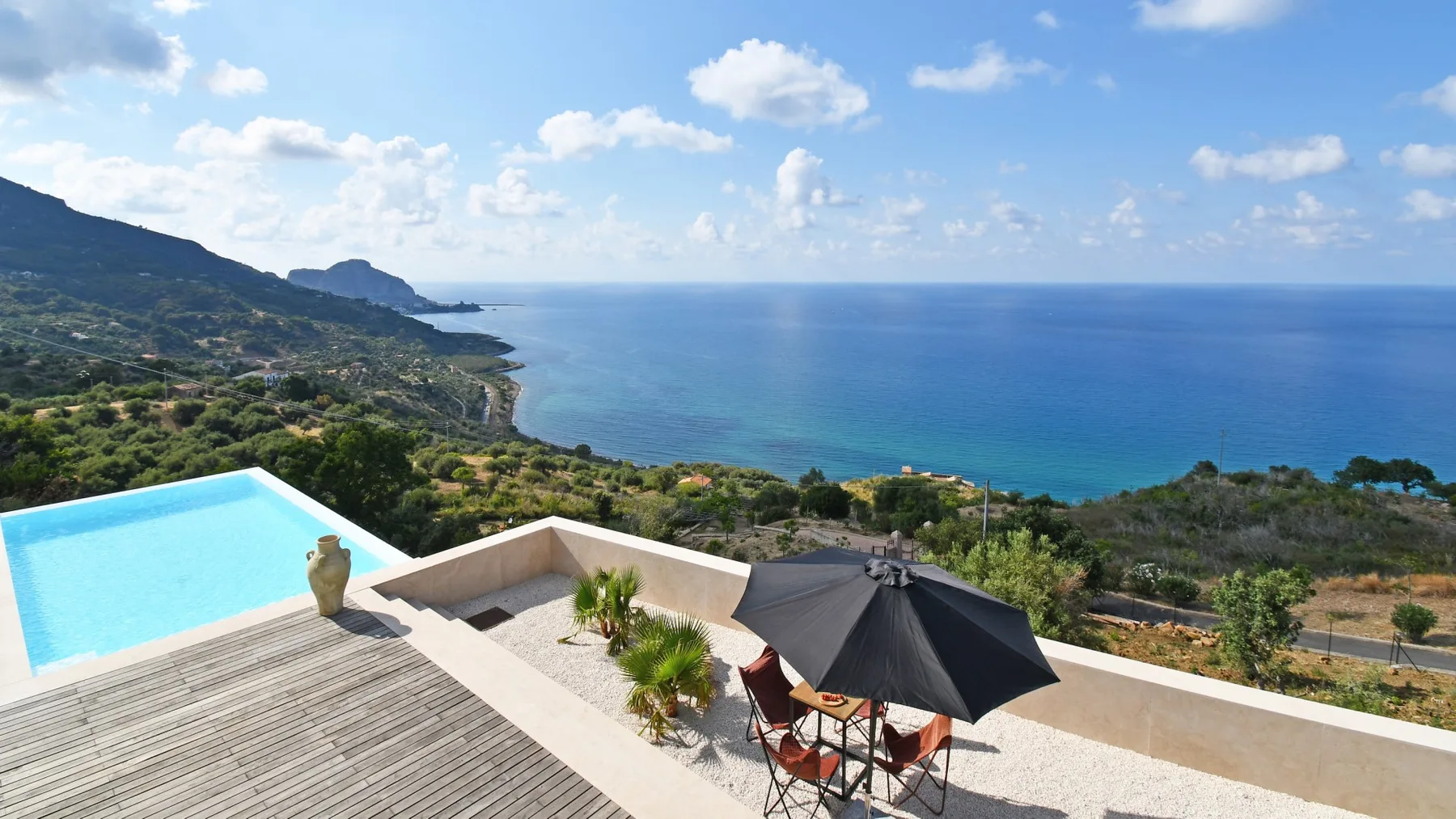 Una terraza en la ladera mira al mar azul, con una pequeña piscina, sillas y una sombrilla negra.