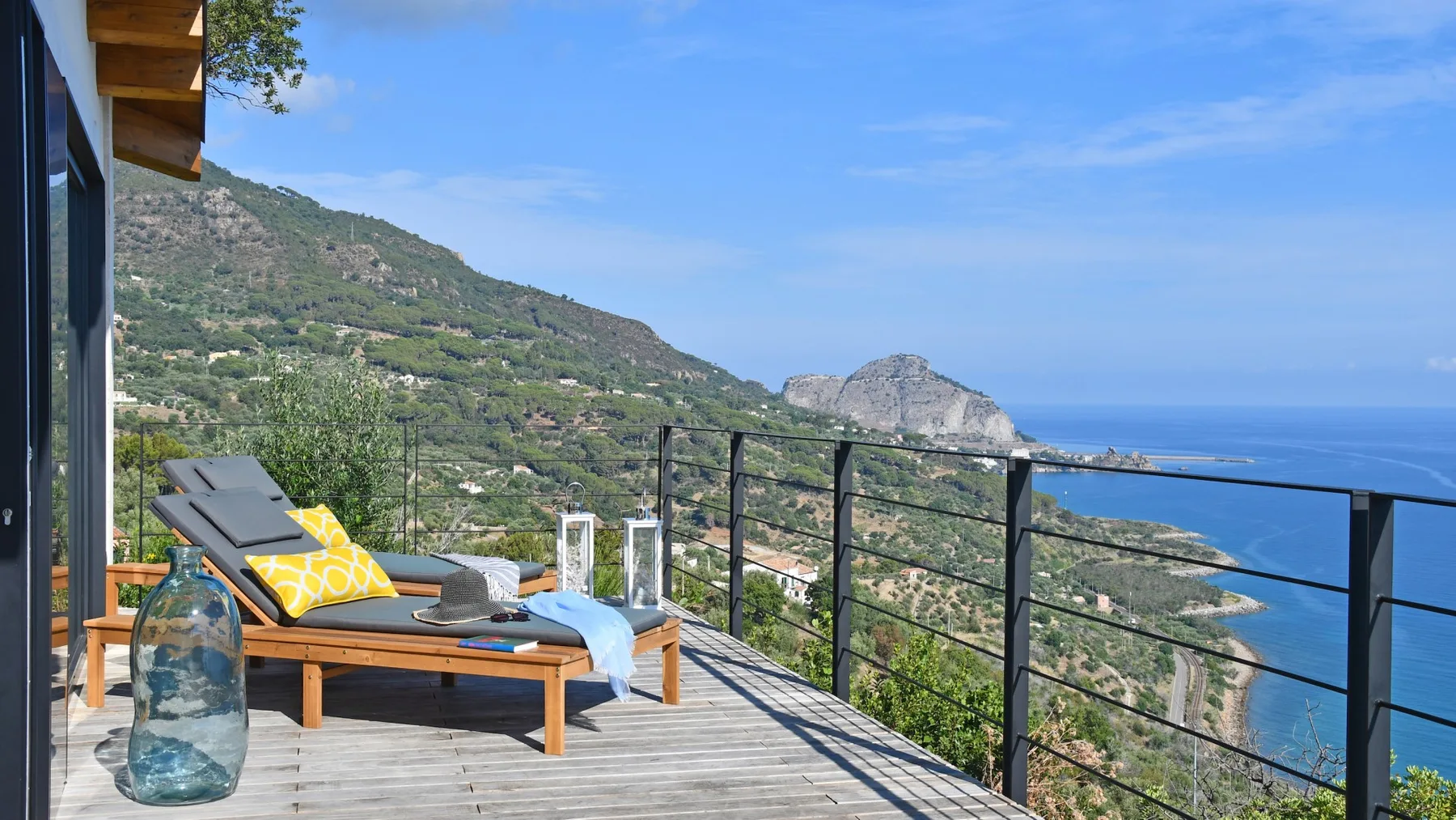 Una terraza de madera con tumbonas y una mesa de vidrio da a una ladera verde y al mar.