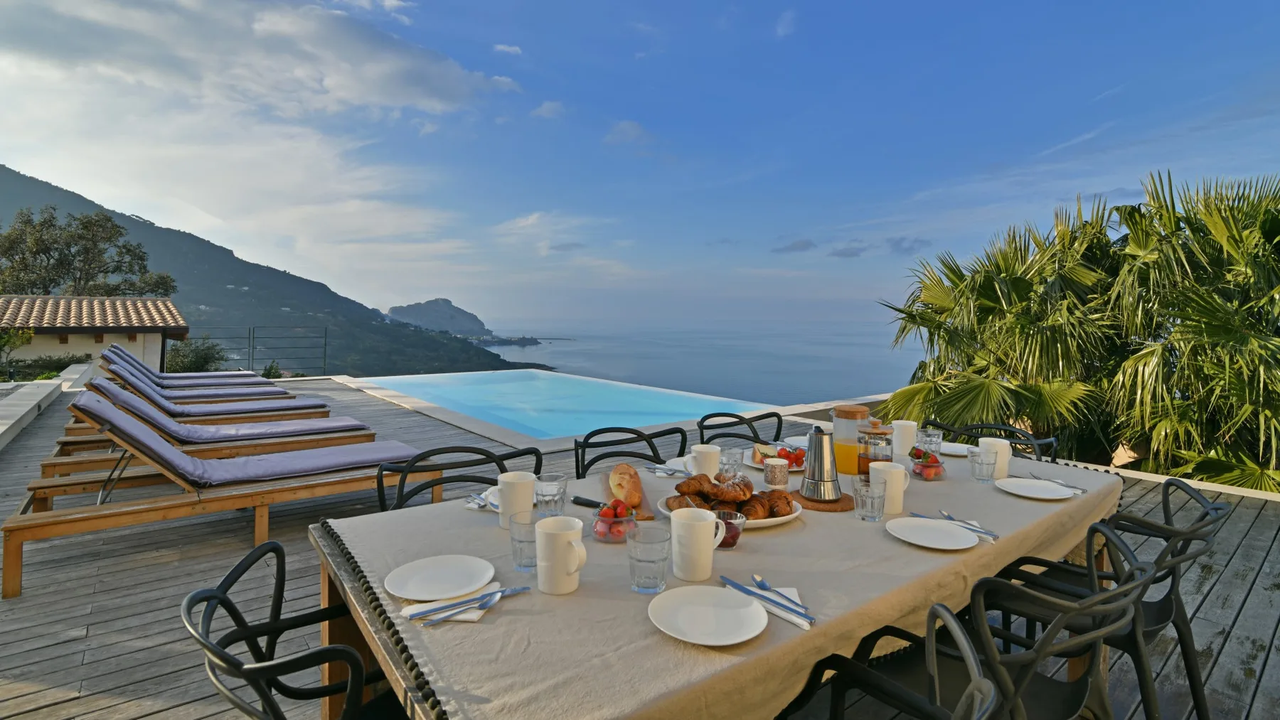 Una mesa al aire libre está preparada con platos y bebidas junto a una piscina, con montañas y el mar al fondo.