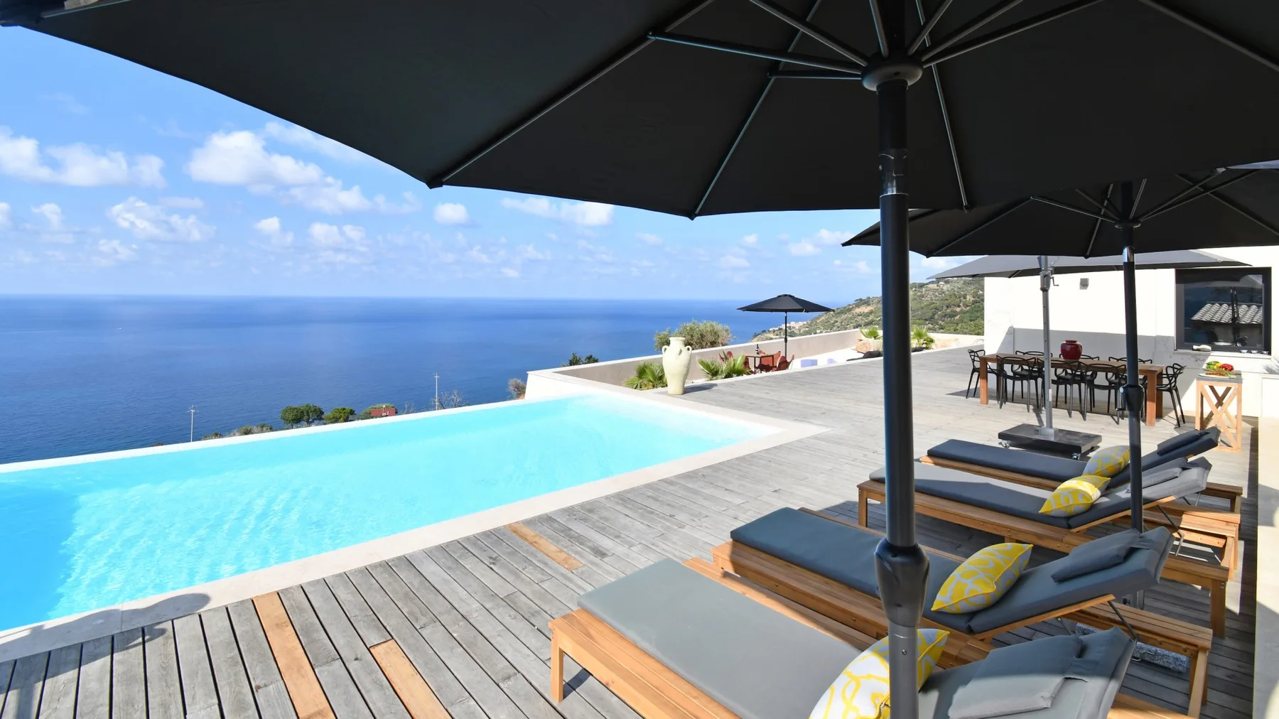 Una piscina junto al mar mira al océano azul, con tumbonas de madera y grandes sombrillas negras en la terraza.