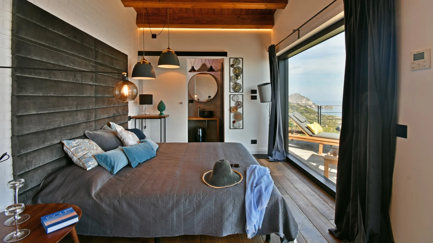 Un dormitorio con una cama grande, lámparas colgantes y puertas de vidrio que dan a un balcón con vistas al mar.