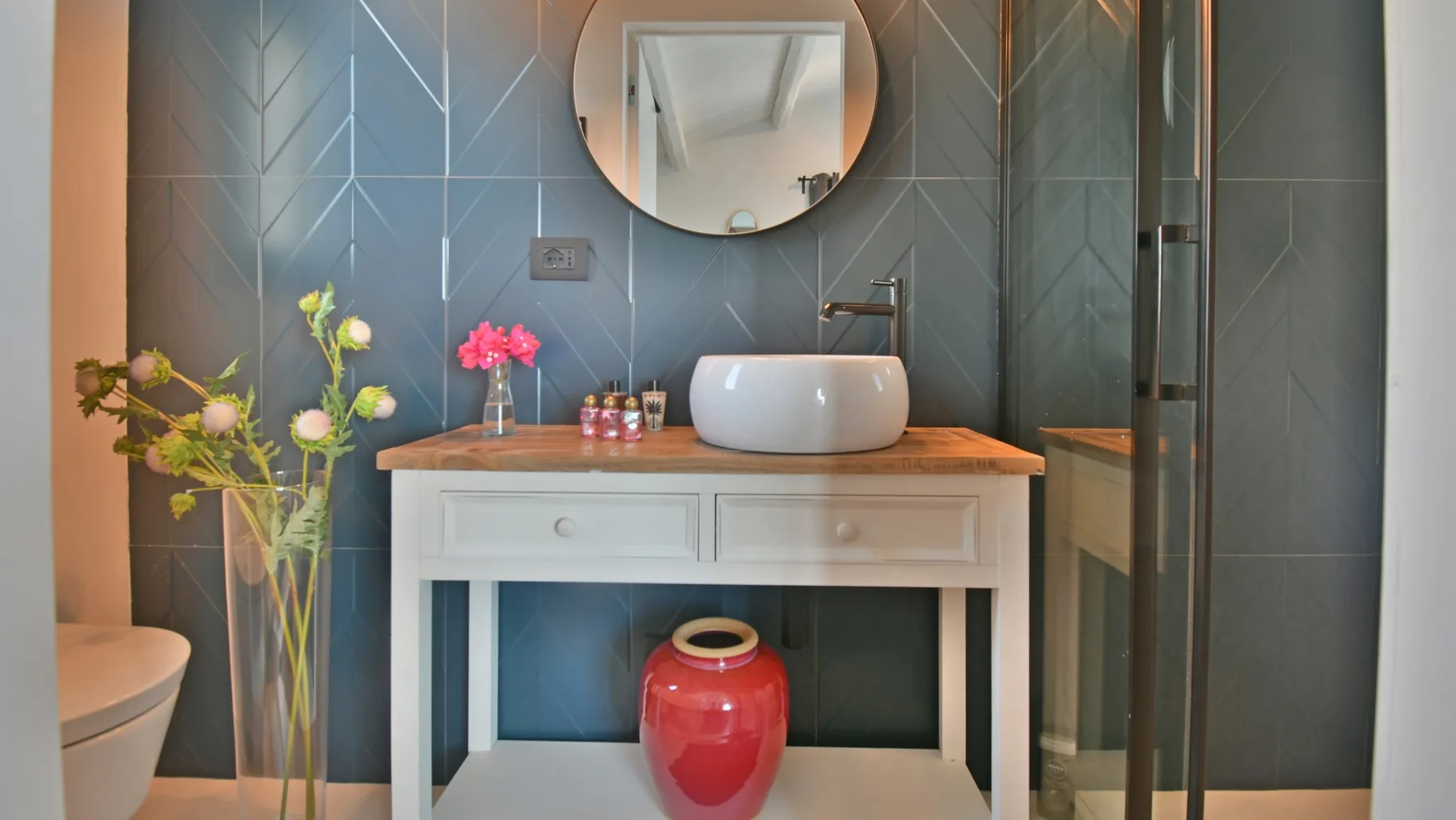 Un tocador de baño con lavabo blanco sobre encimera, espejo redondo, azulejos azul grisáceo y un taburete rojo.