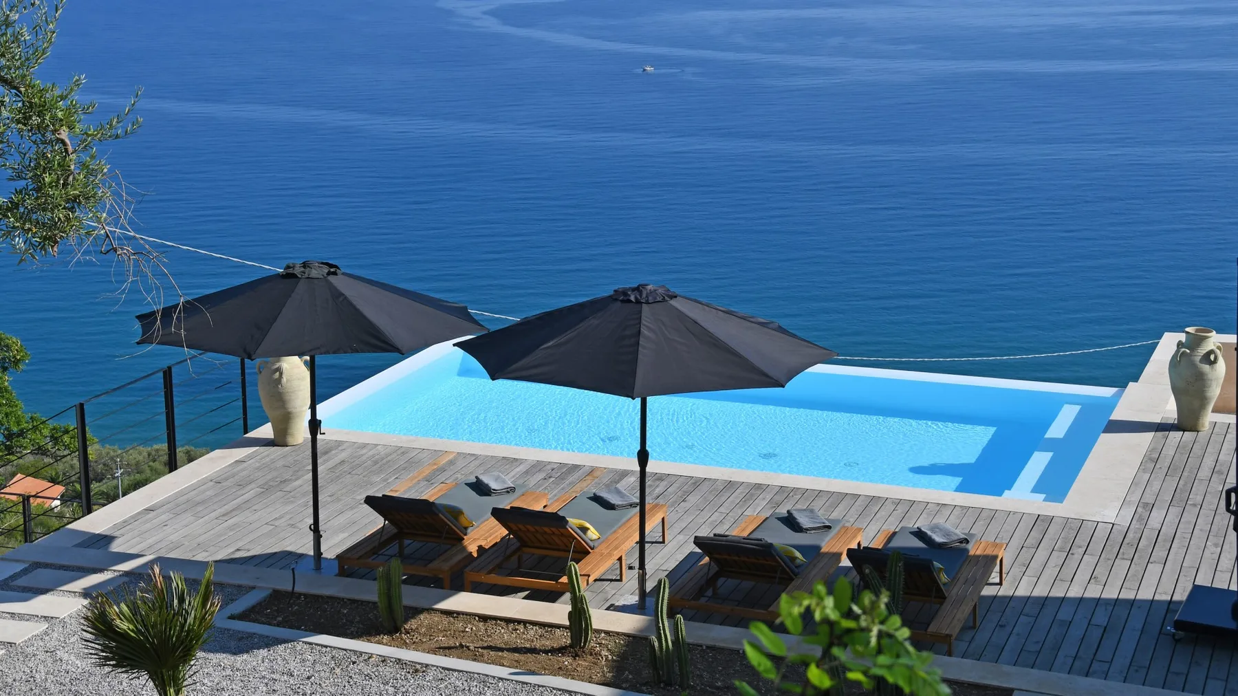 Terraza junto a una piscina al aire libre, con sombrillas oscuras y tumbonas frente al mar.