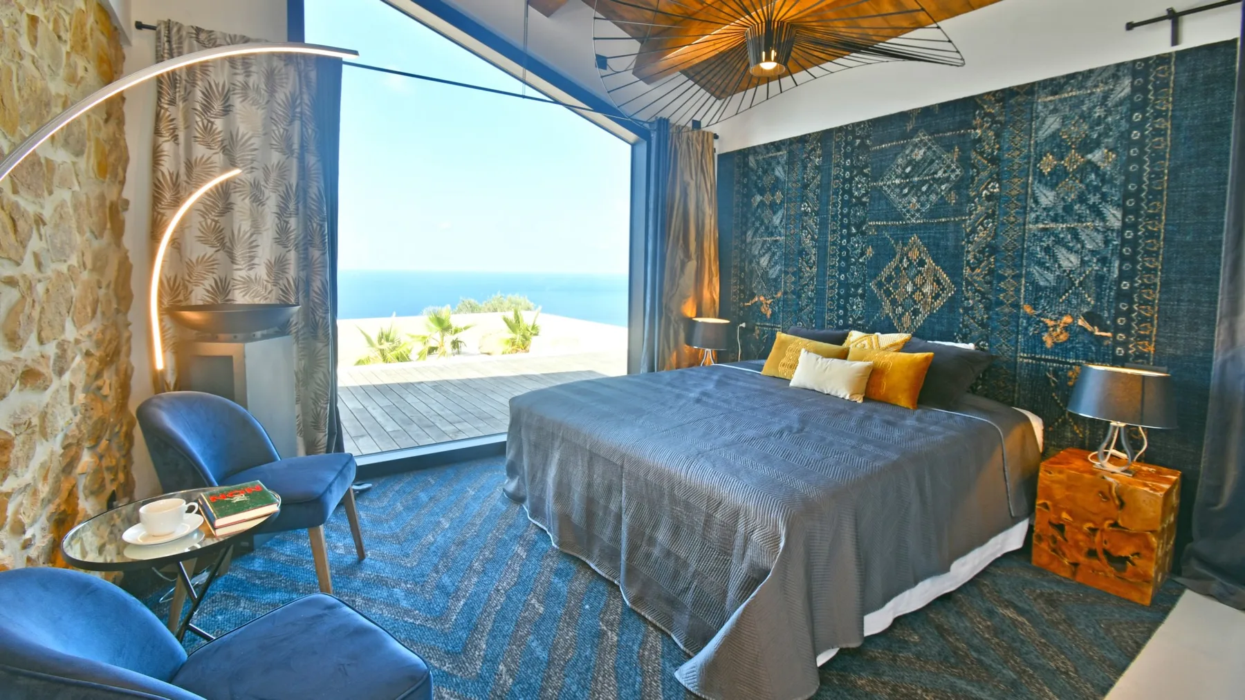 Un dormitorio con una gran cama gris, paredes estampadas azules y una vista abierta al mar.