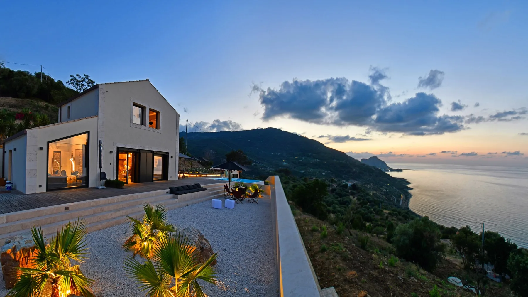 Una casa blanca moderna y una terraza con palmeras miran hacia una ladera costera verde y un mar tranquilo al atardecer.