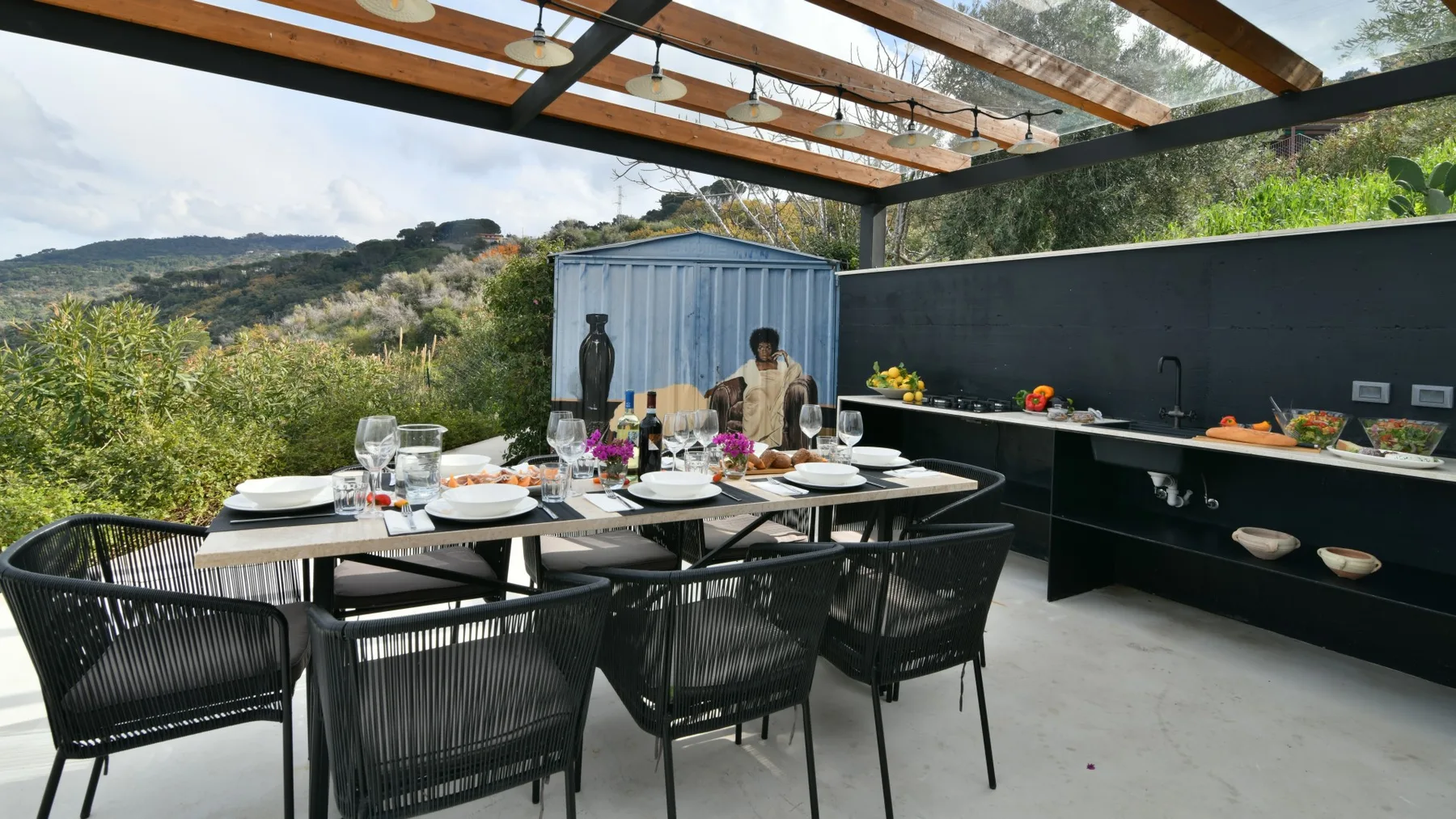 Una zona de comedor al aire libre tiene una mesa larga puesta, sillas tejidas y una cocina exterior cubierta.