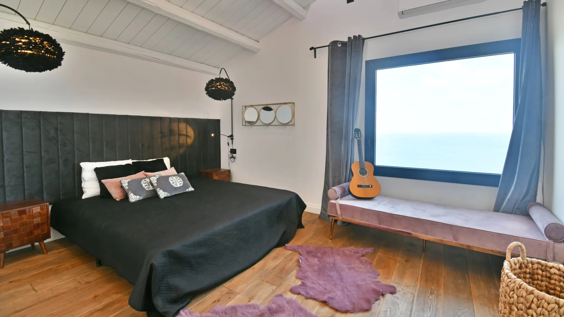 Un dormitorio con una cama grande y oscura, cortinas grises, una guitarra y una ventana amplia.