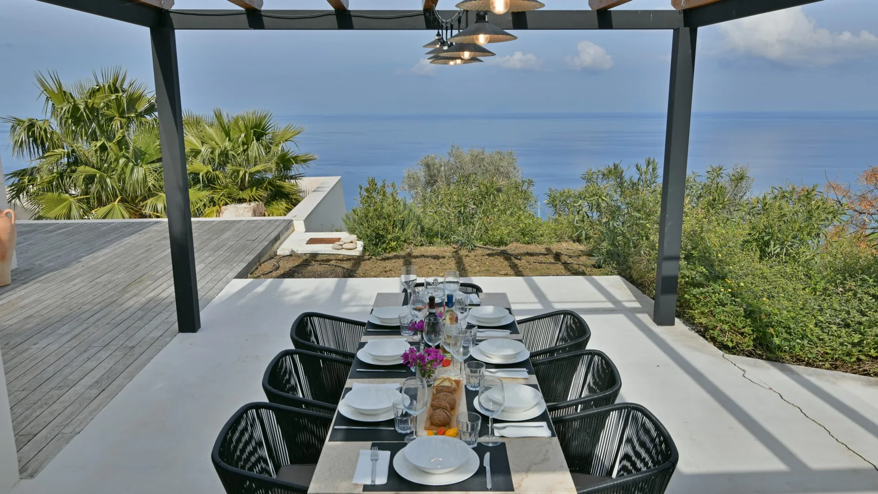 Una larga mesa está preparada bajo una pérgola en una terraza soleada con vistas al mar.