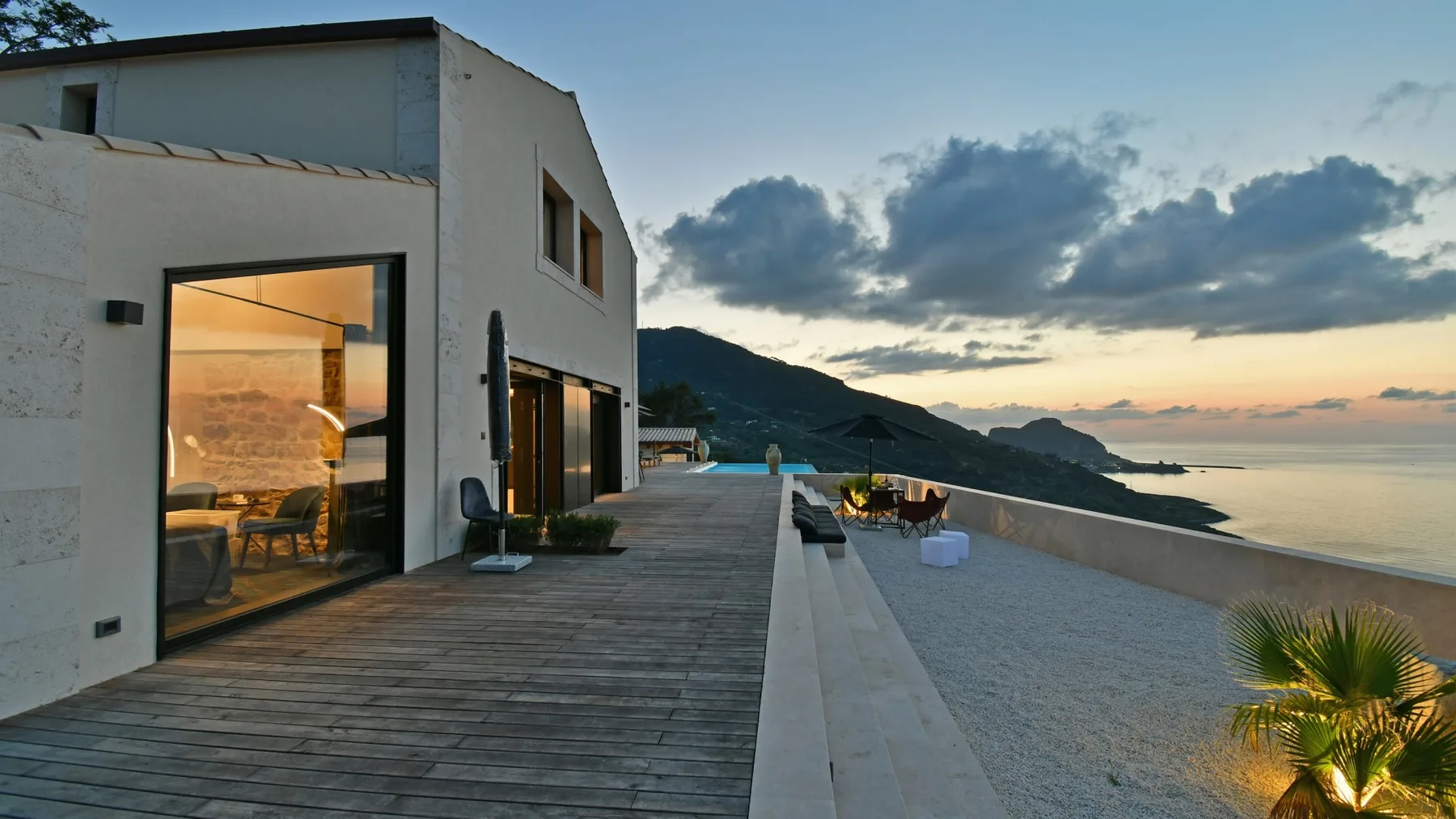 Una casa blanca moderna se abre a una terraza de madera con vistas al mar al atardecer.