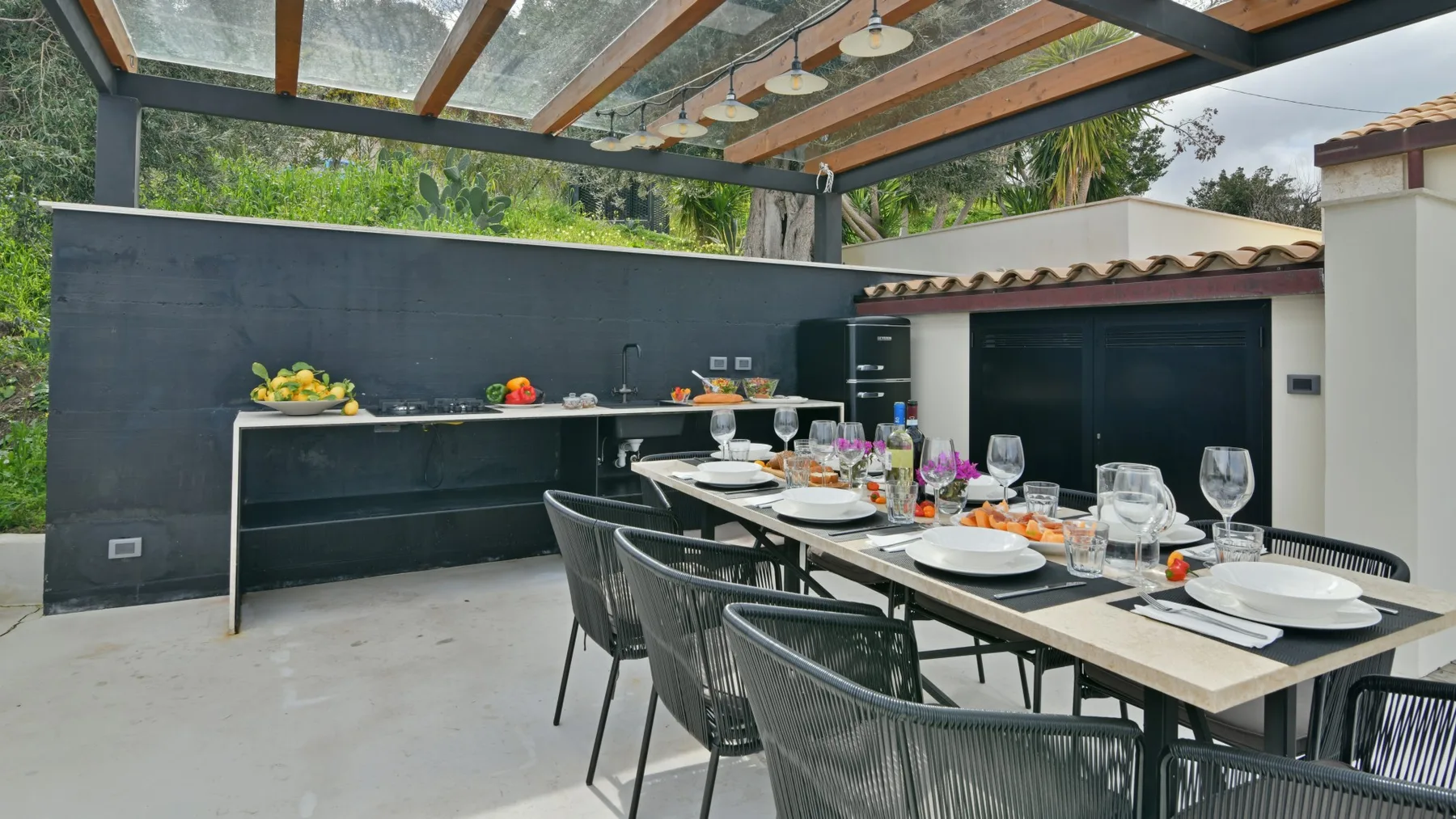 Una zona de comedor exterior cubierta tiene una mesa larga puesta con platos y copas junto a una encimera oscura.