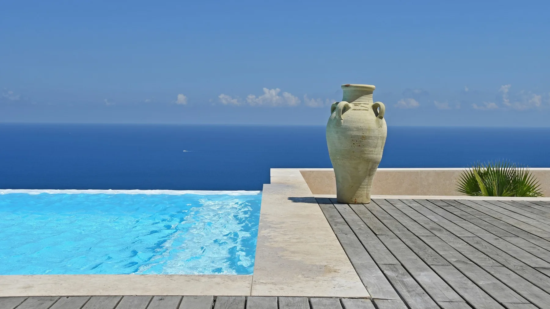 Una piscina azul se une a una terraza de madera, con un gran jarrón de cerámica y el mar al fondo.