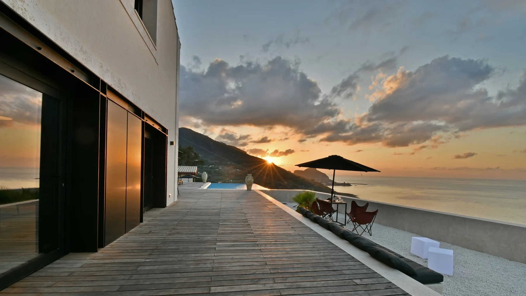 Una terraza de madera junto a un edificio moderno da a la playa, con sillas bajo una sombrilla al atardecer.