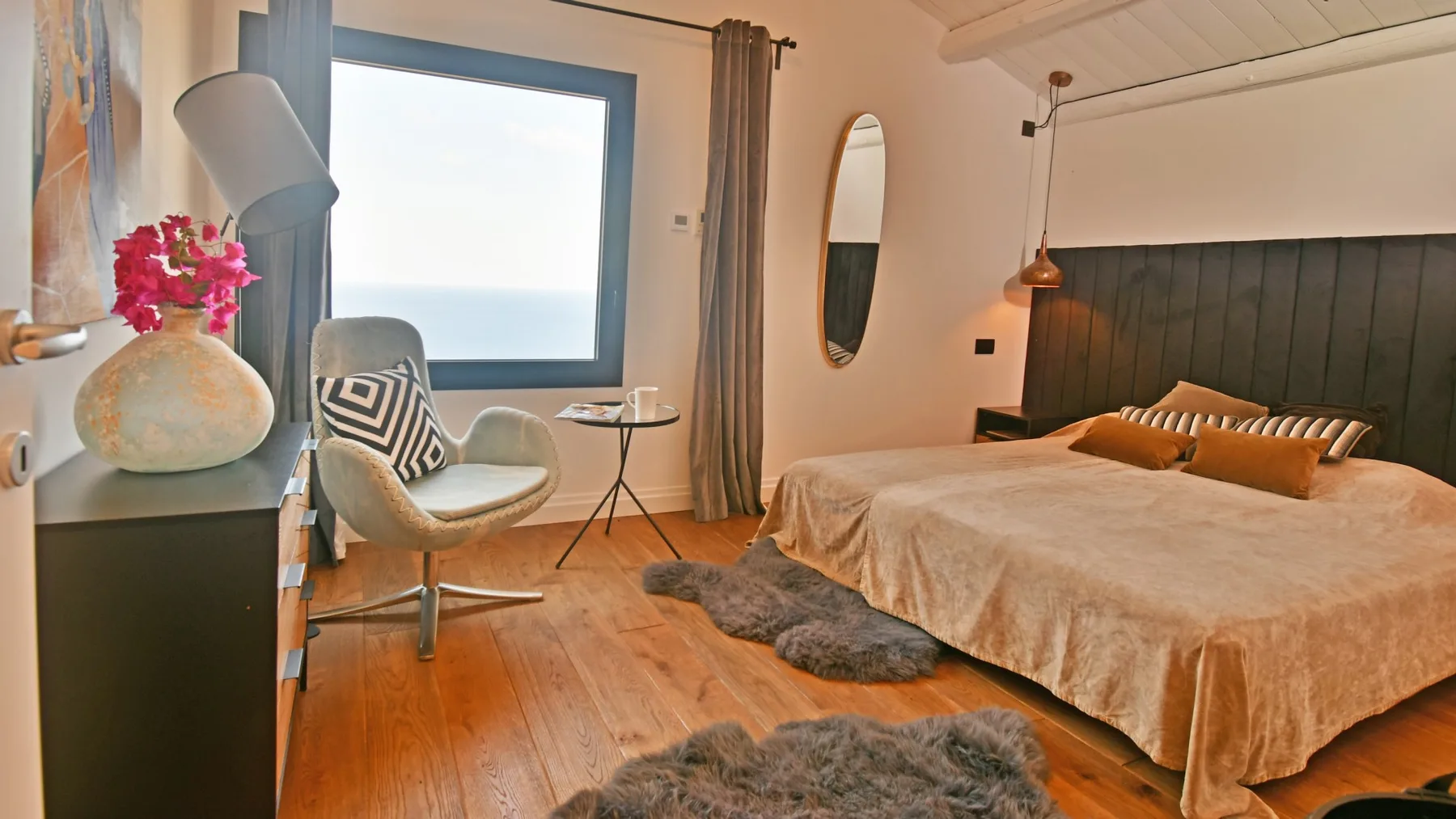 Un dormitorio con una cama grande, un sillón tapizado, una lámpara y una amplia ventana con vistas al agua.
