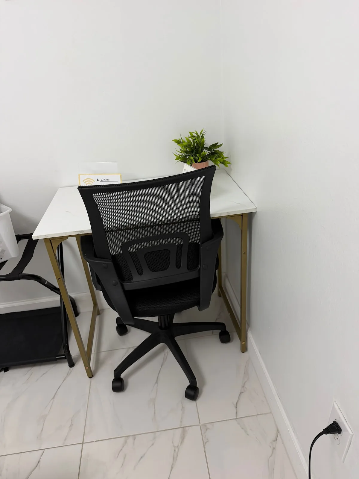 A small office desk and chair sit against a plain wall, with a potted plant on the desk.