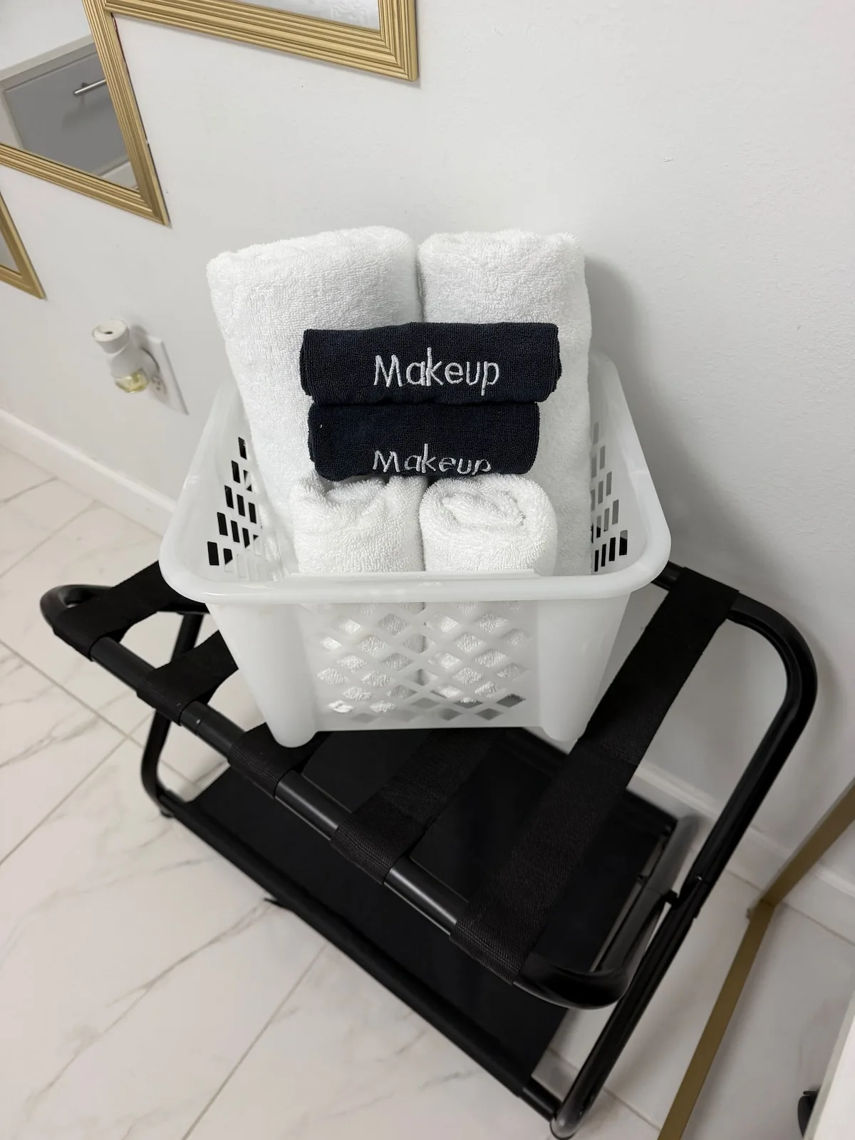 A basket of folded white towels and black makeup cloths sits on a luggage rack.