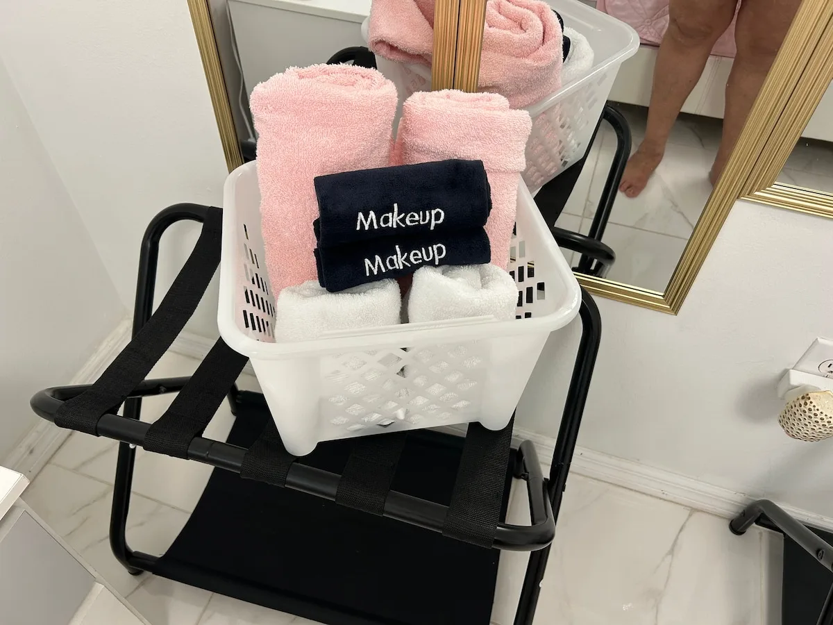 A white laundry basket holding folded towels sits on a black folding chair.