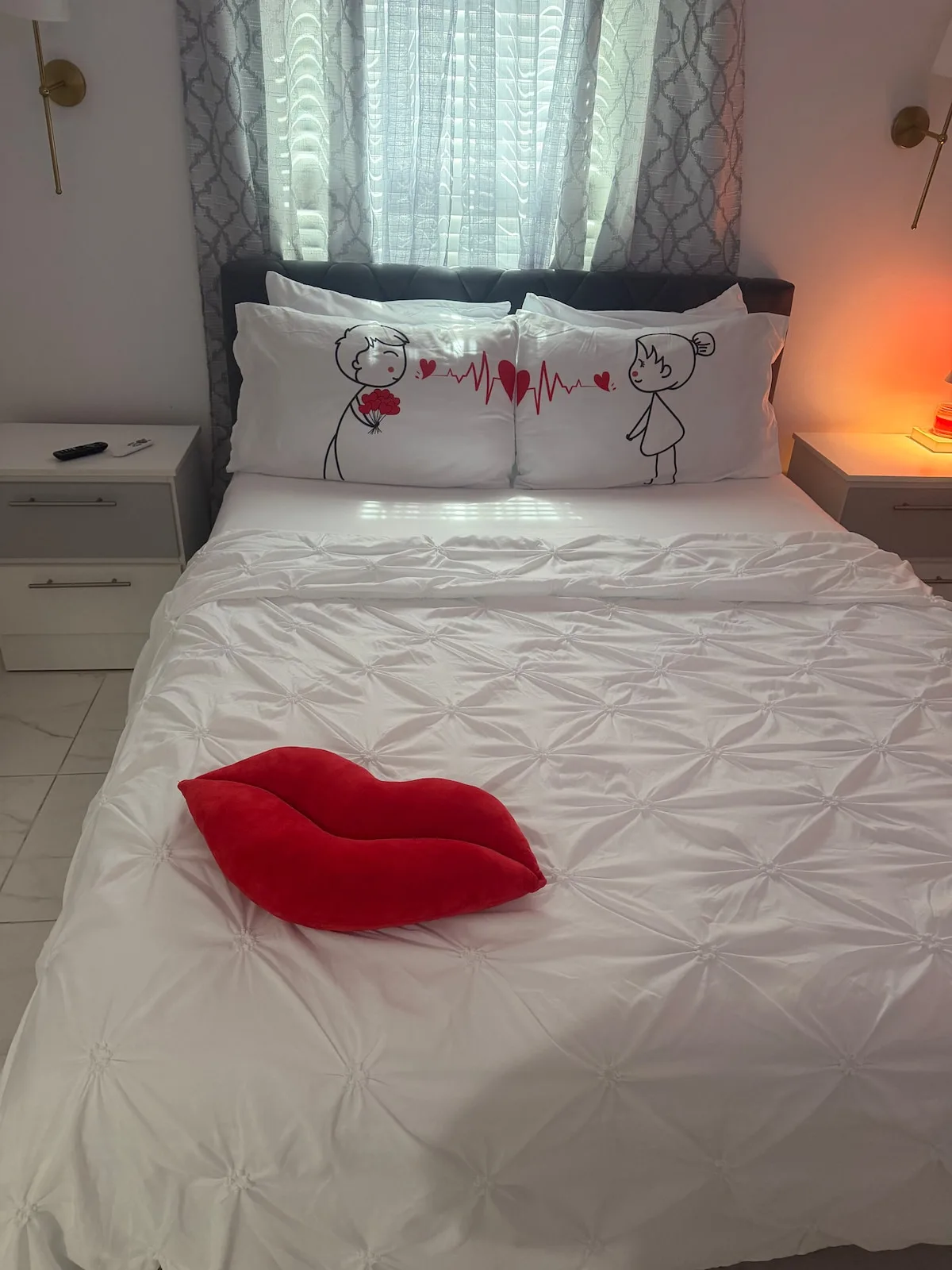 A bedroom with white bedding and a red heart-shaped cushion on the bed.