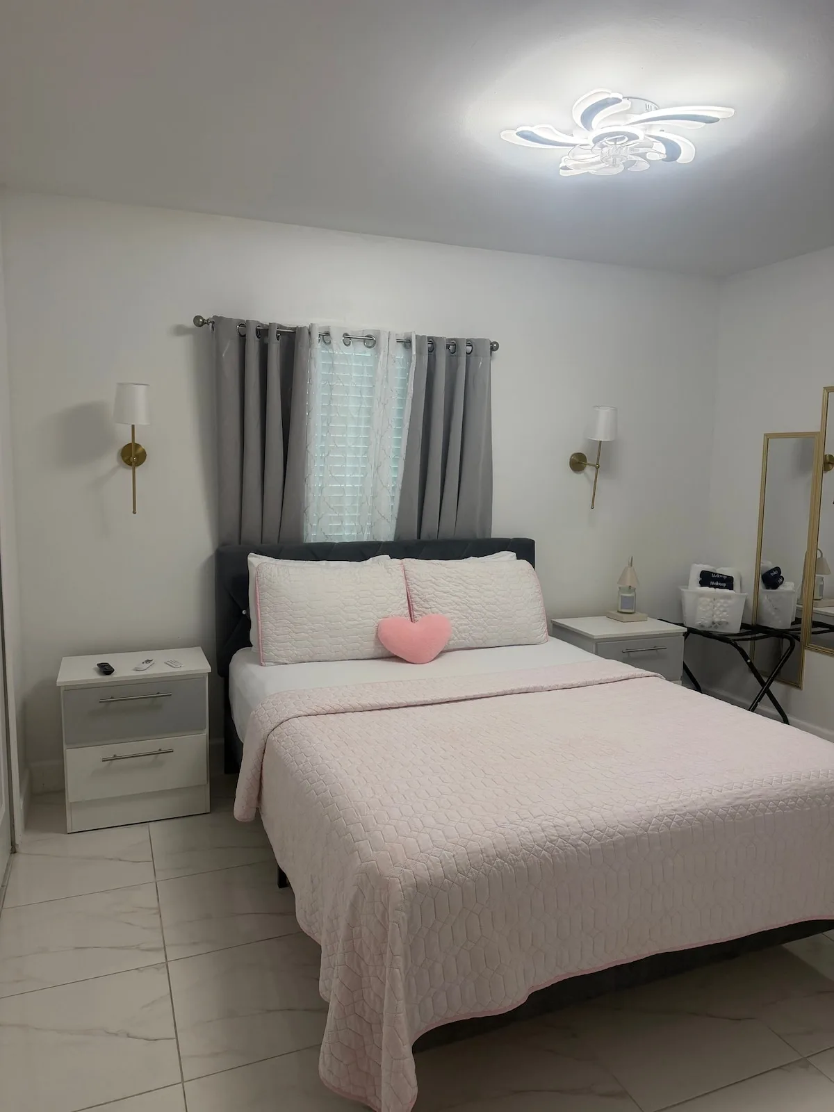 A bedroom with a bed dressed in pale pink bedding beneath a curtained window.