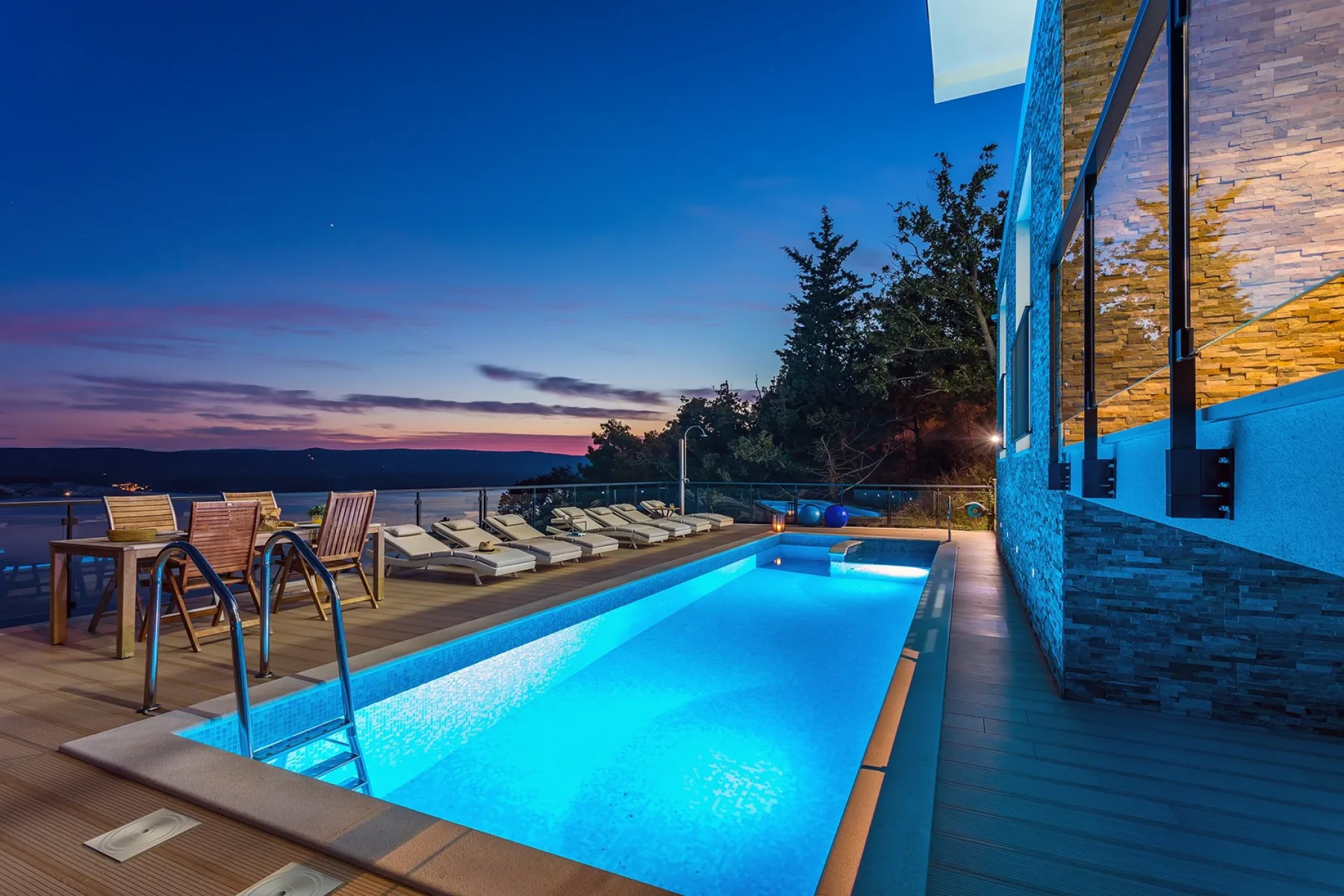 A swimming pool beside a modern building at dusk, with loungers on the deck.
