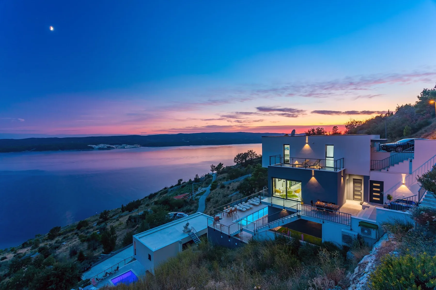 A modern house sits on a hillside overlooking a body of water at sunset.